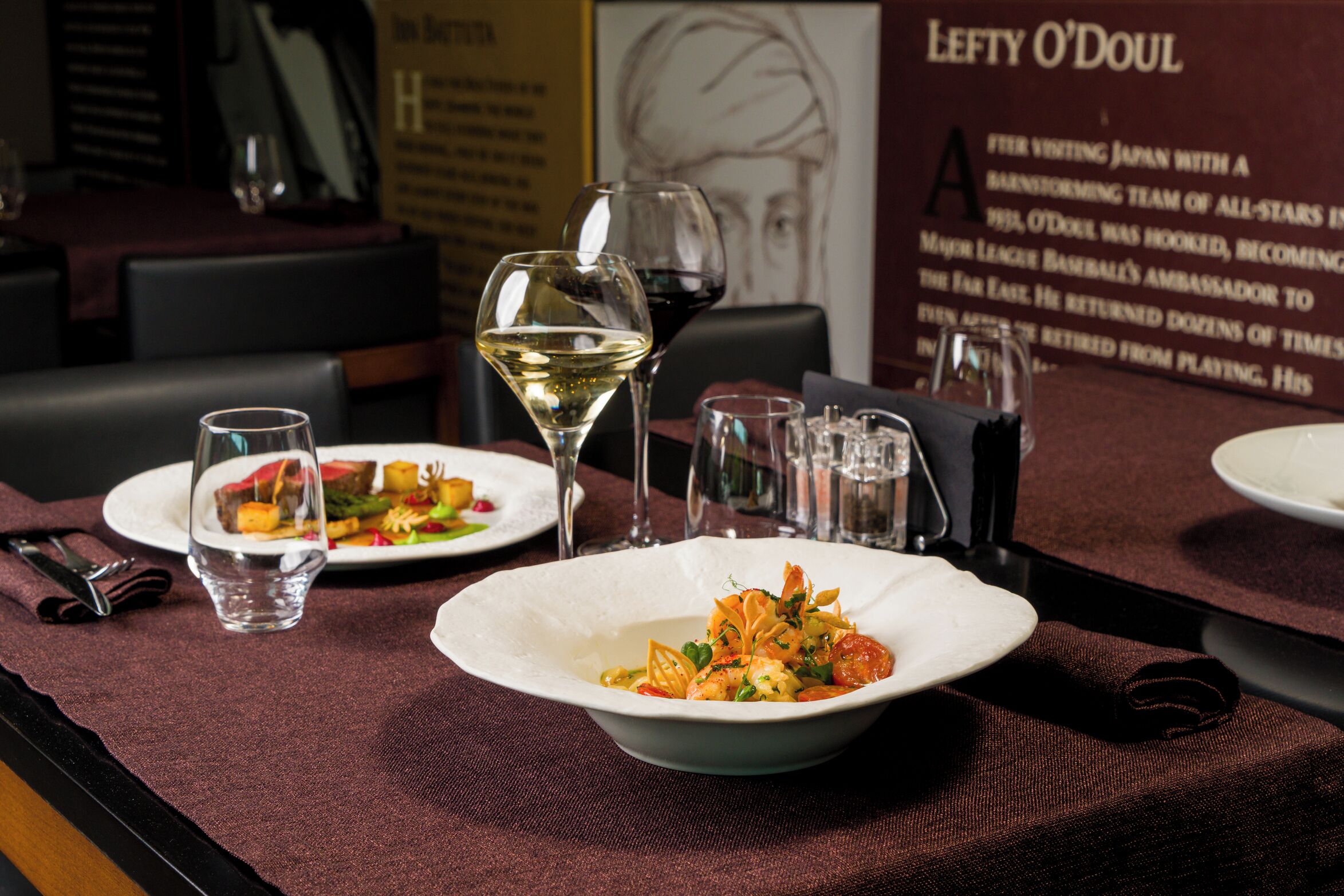 A dining table with plates of food, glasses of water and wine, and a menu board in the background.