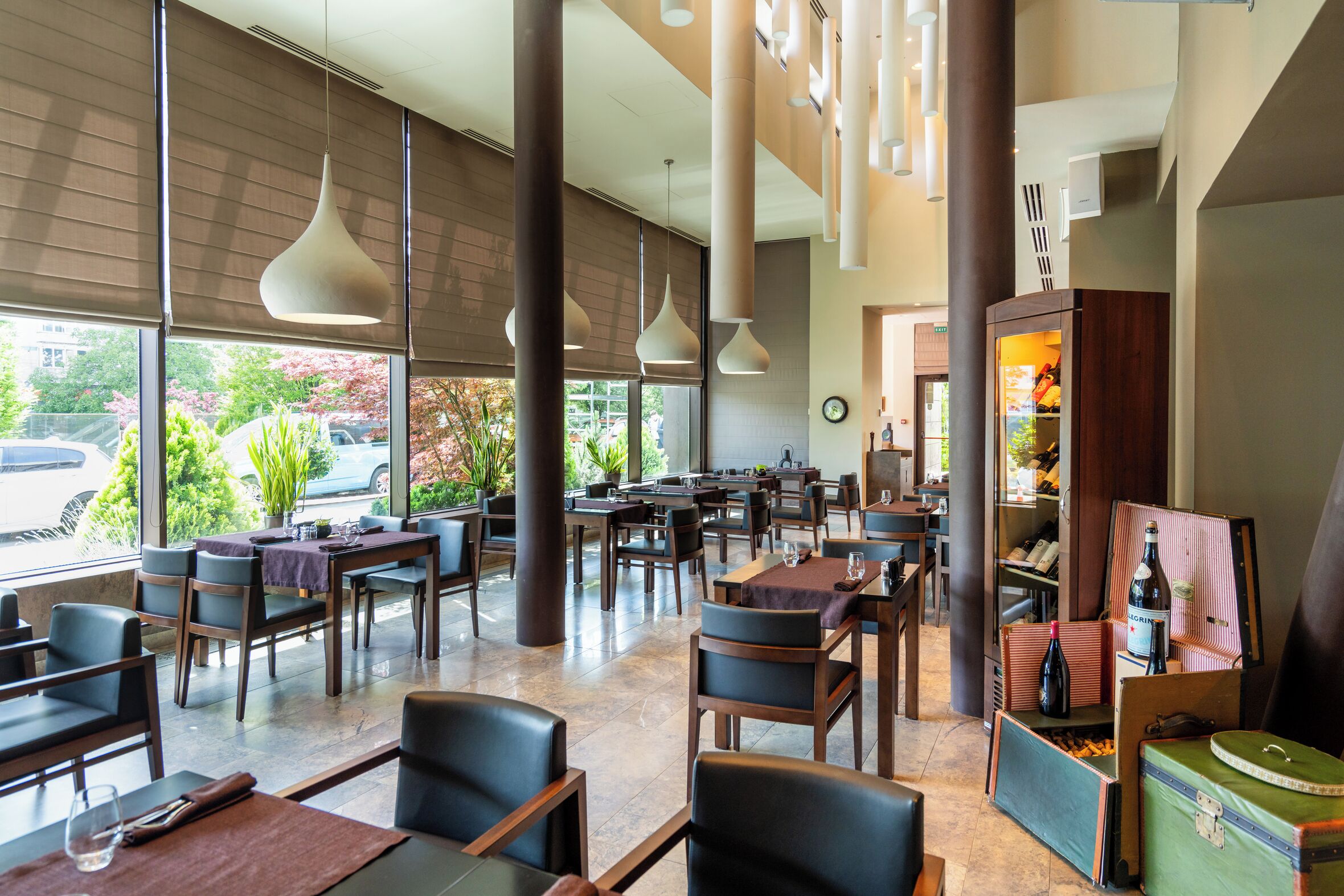 A spacious restaurant with high ceilings, large windows, and modern pendant lighting. Tables are arranged for dining, and there are wine bottles and glasses on display.