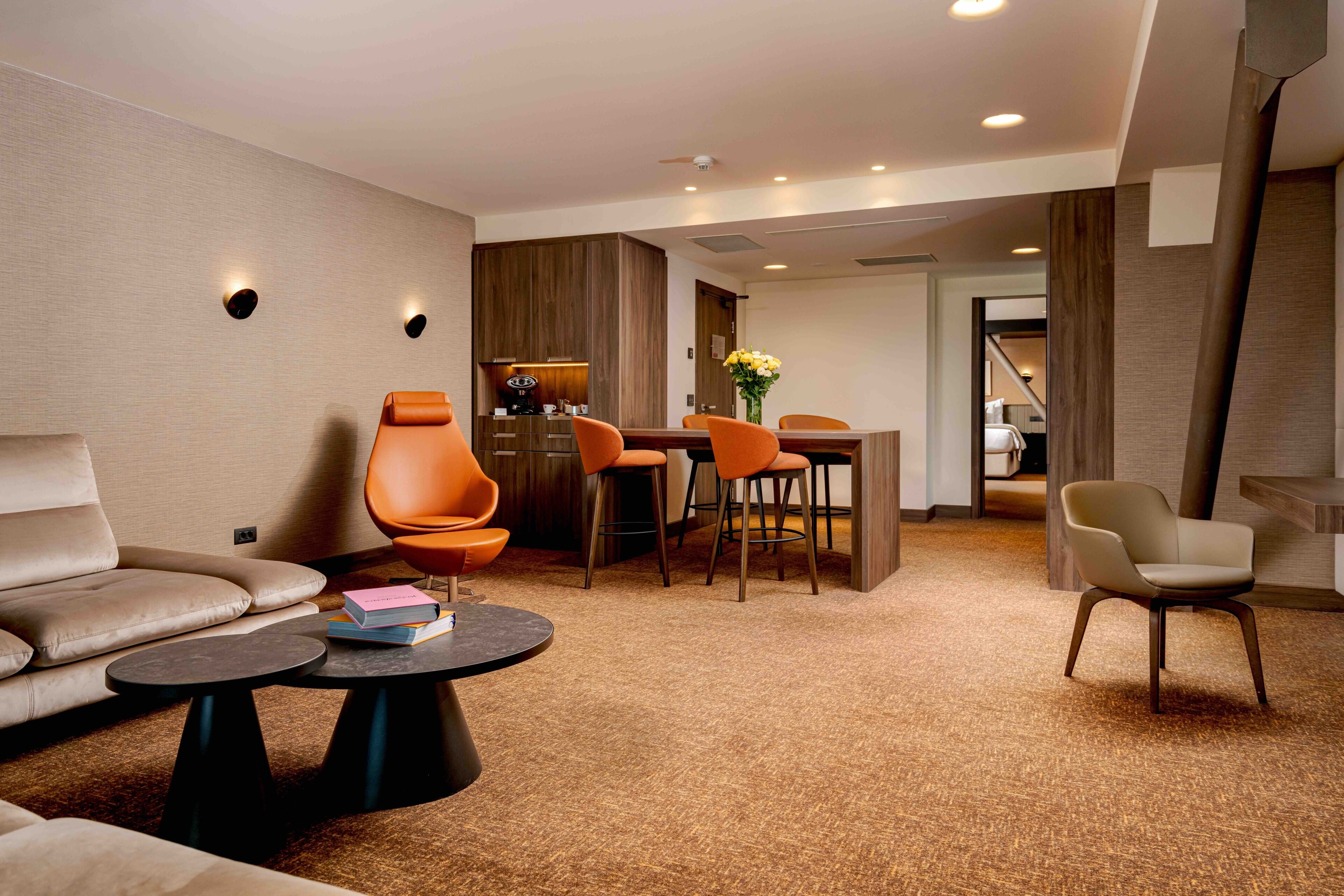 A modern hotel room with a sofa, a round coffee table, a dining area with chairs, and a kitchen area.