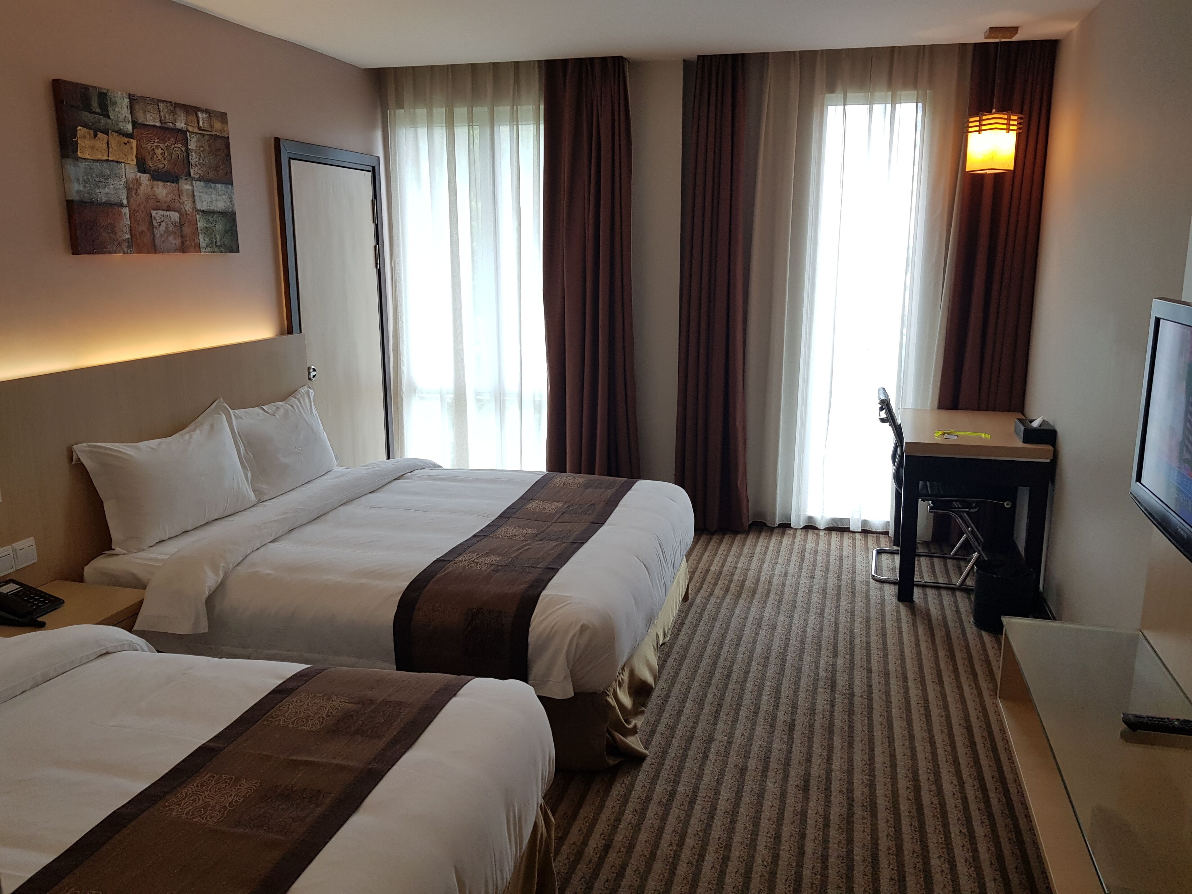 A hotel room with two beds, a desk, a television, and a large window with curtains.
