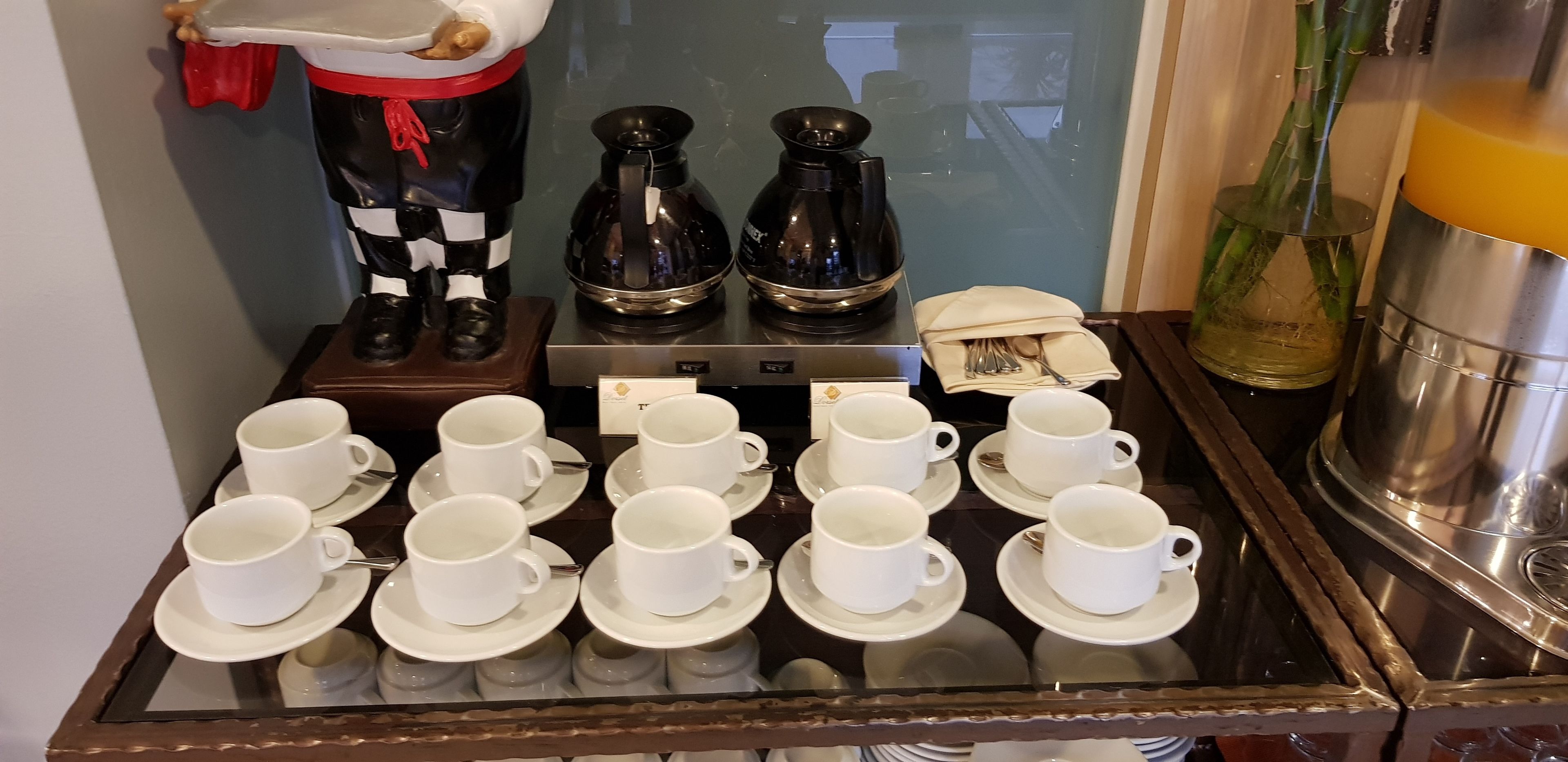 A tray with multiple white cups and saucers, a coffee maker, and a decorative figure.