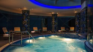 Piscina interior com teto de vidro, rodeada por espreguiçadeiras e pilares estampados.