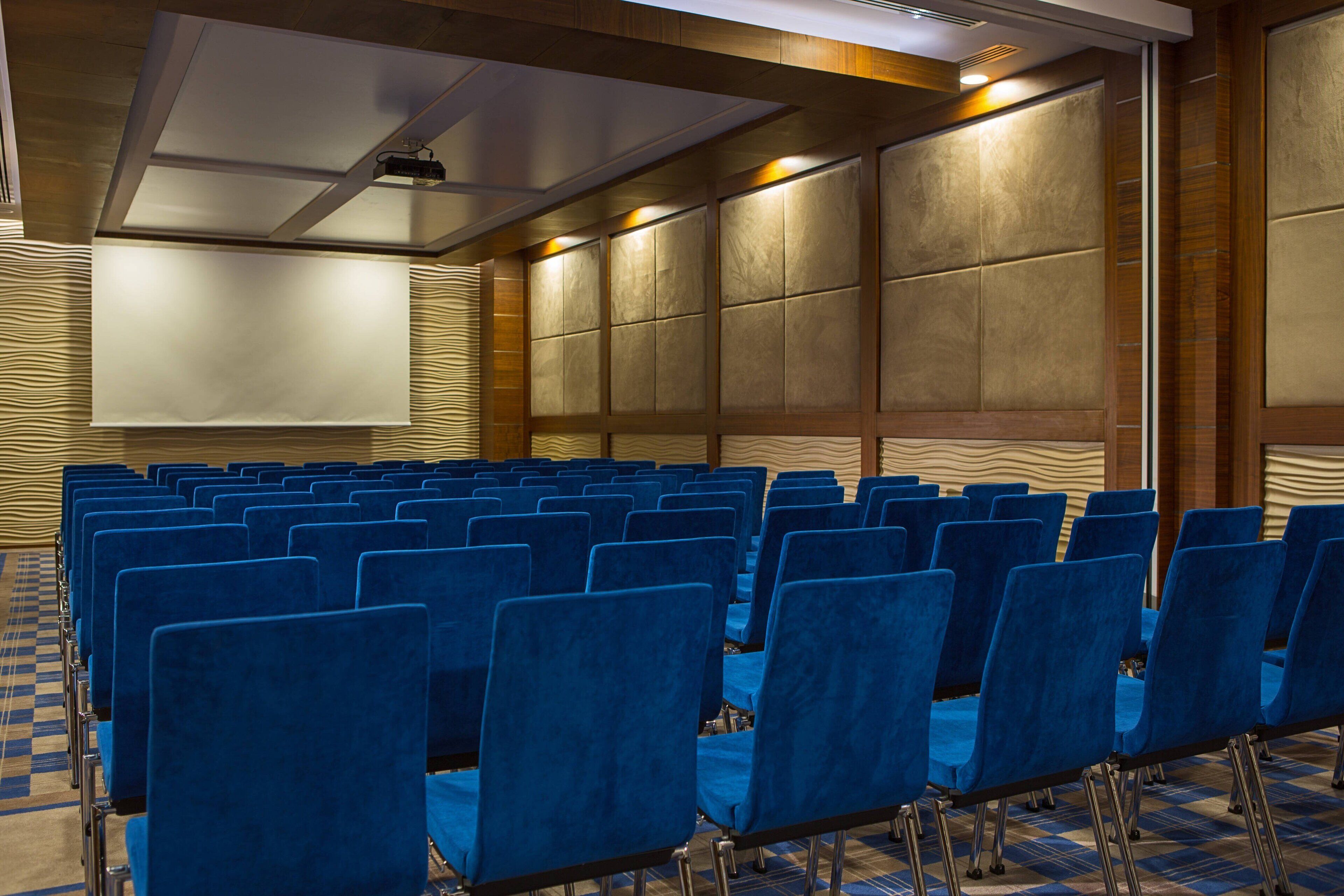 Une salle de conférence avec des rangées de chaises bleues, un grand écran et une moquette à motifs.