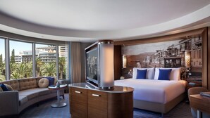 Quarto de hotel moderno com uma cama curva, um sofá, uma TV e uma janela ampla com vista para a cidade.