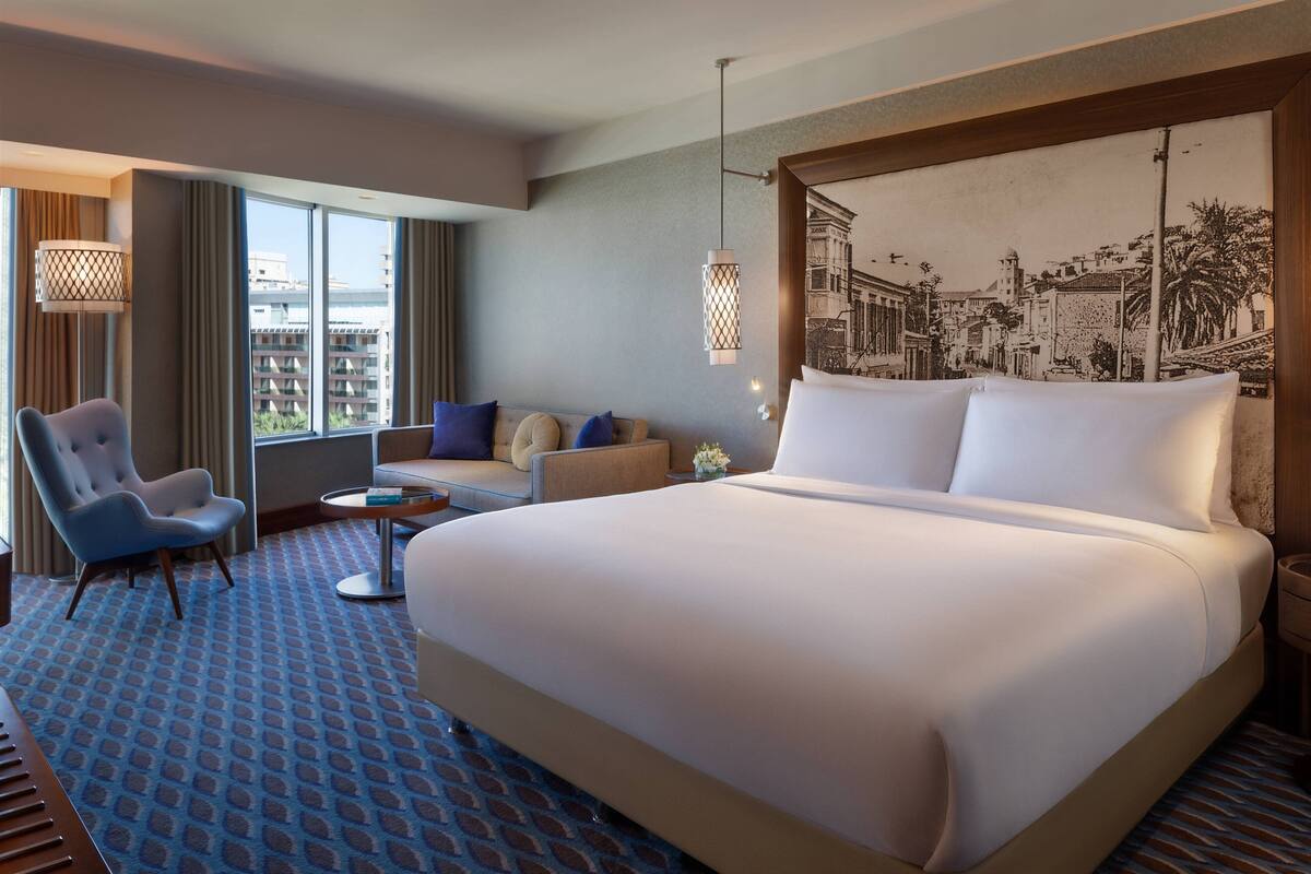 A hotel room with a large bed, a sofa, a chair, a small table, and a large window offering a city view.