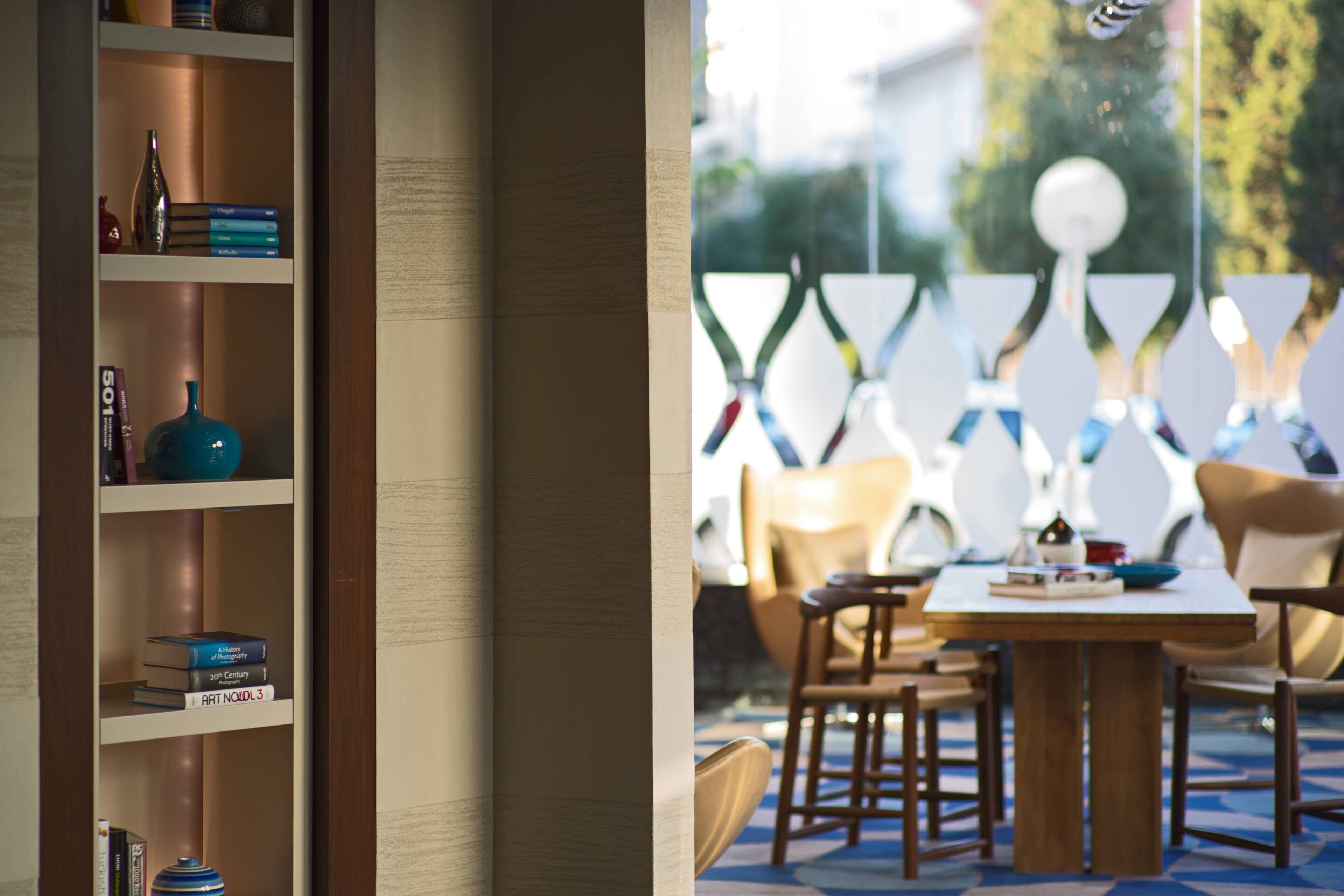 A modern interior with a wooden bookshelf, a dining area with a table and chairs, and a view of the outdoors through a window.