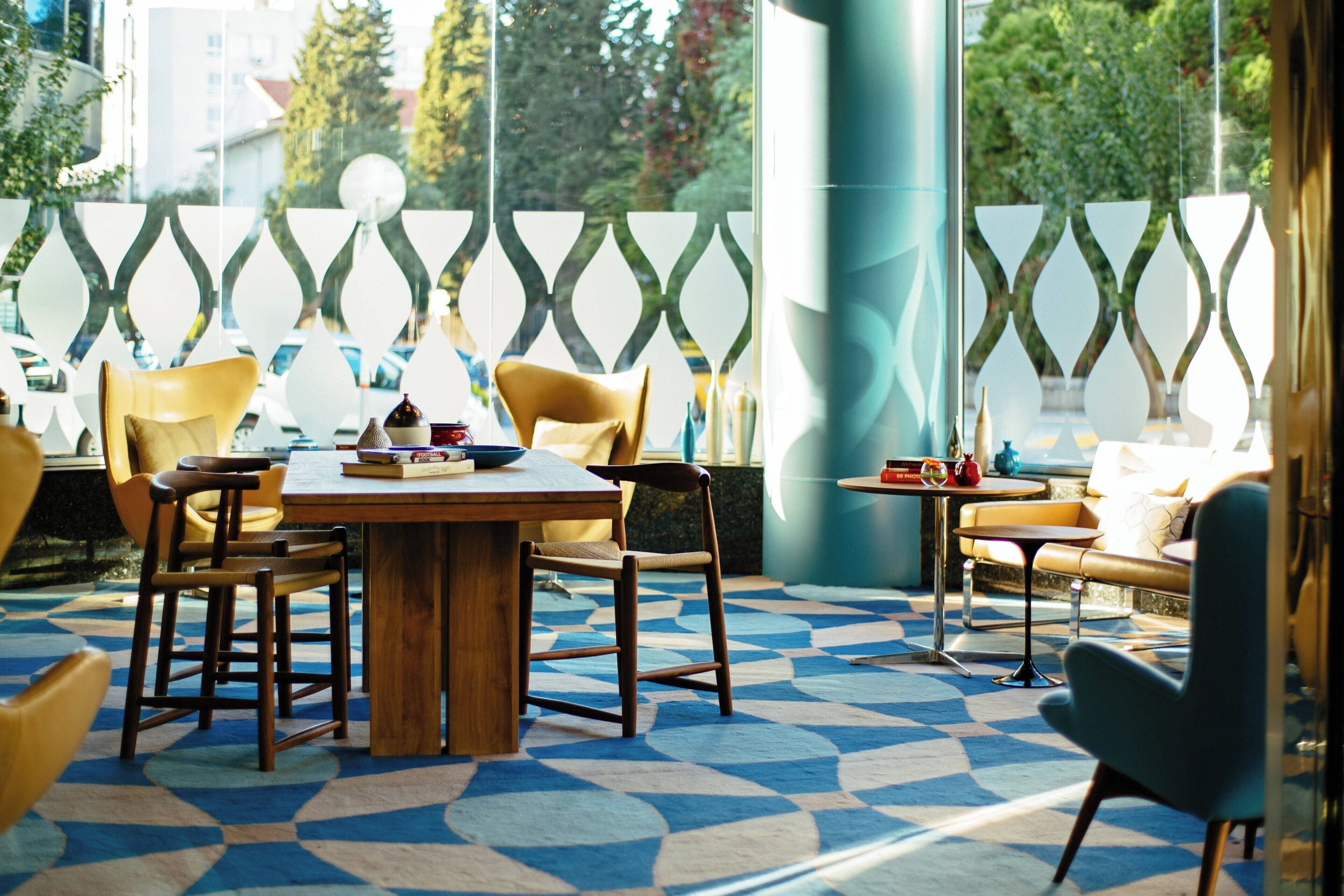 Ein modernes Interieur mit einem Holztisch, gelben Stühlen, einem blau gemusterten Boden und Blick durch eine Fensterwand mit Muster.