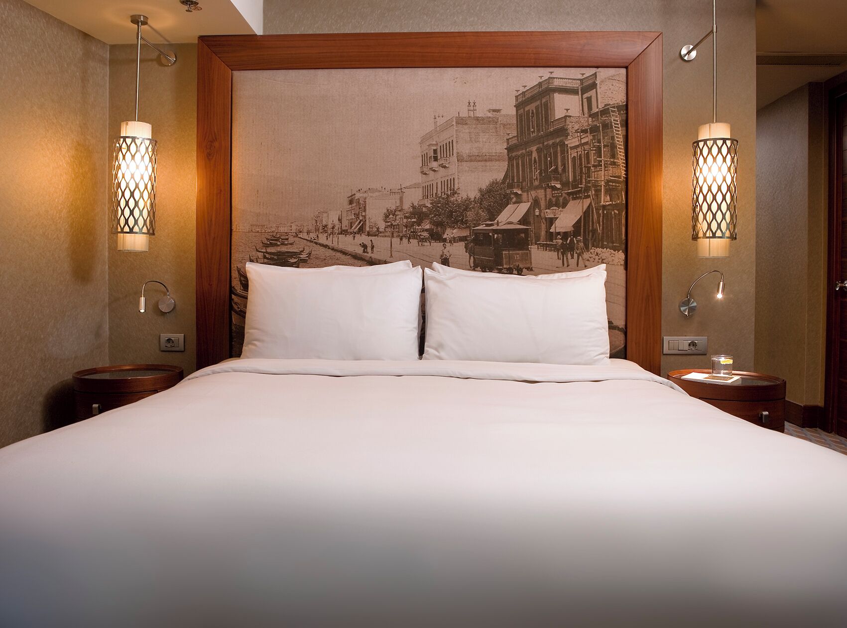 A hotel room with a large bed, two bedside lamps, a wooden headboard, and a vintage cityscape wall art.