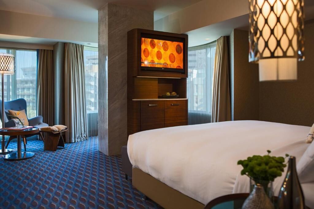 A hotel room with a large bed, a television mounted on a wooden cabinet, a desk with a chair, and a large window with curtains.