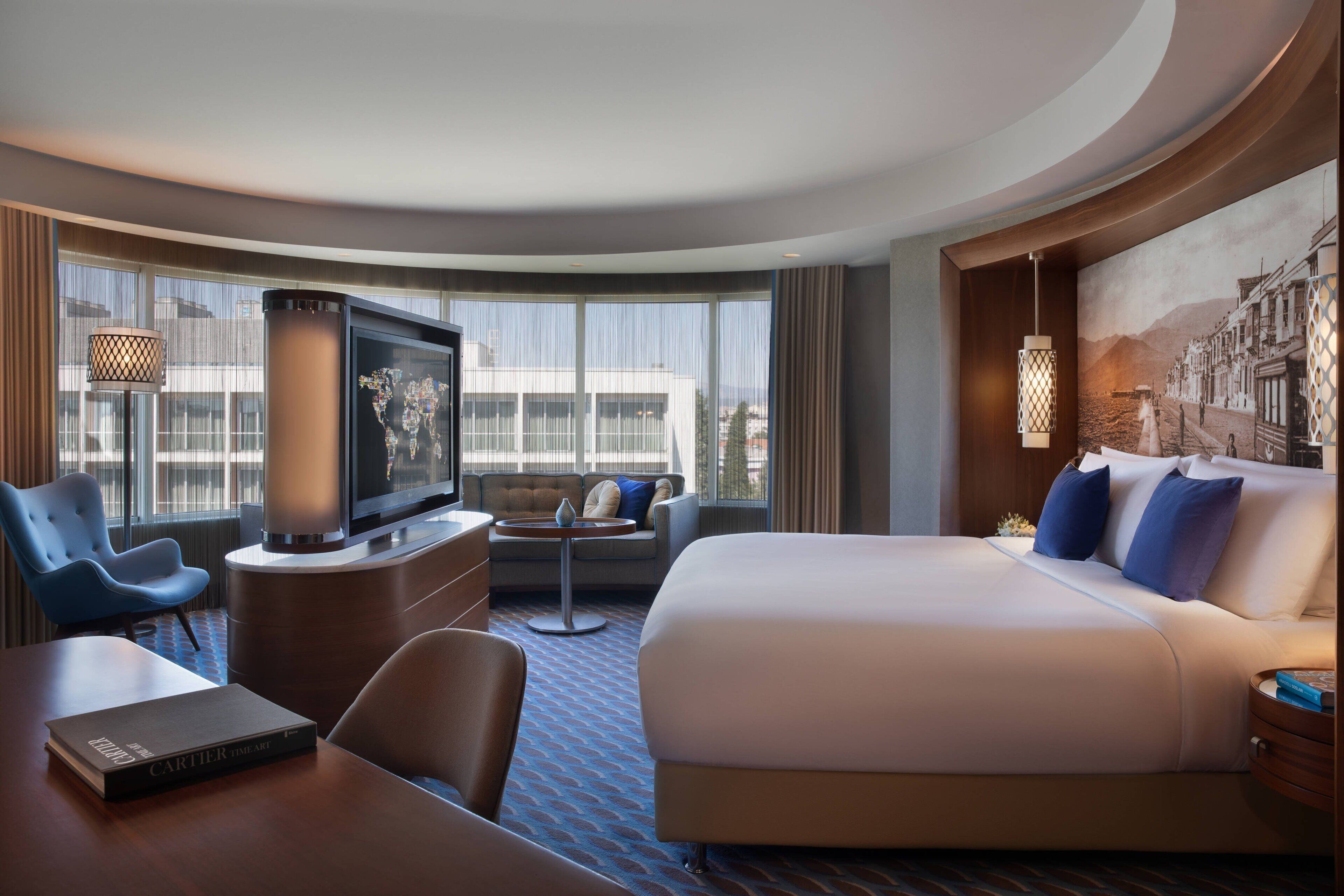 A hotel room with a large bed, a desk with a chair, a television, and a view of a cityscape through large windows.