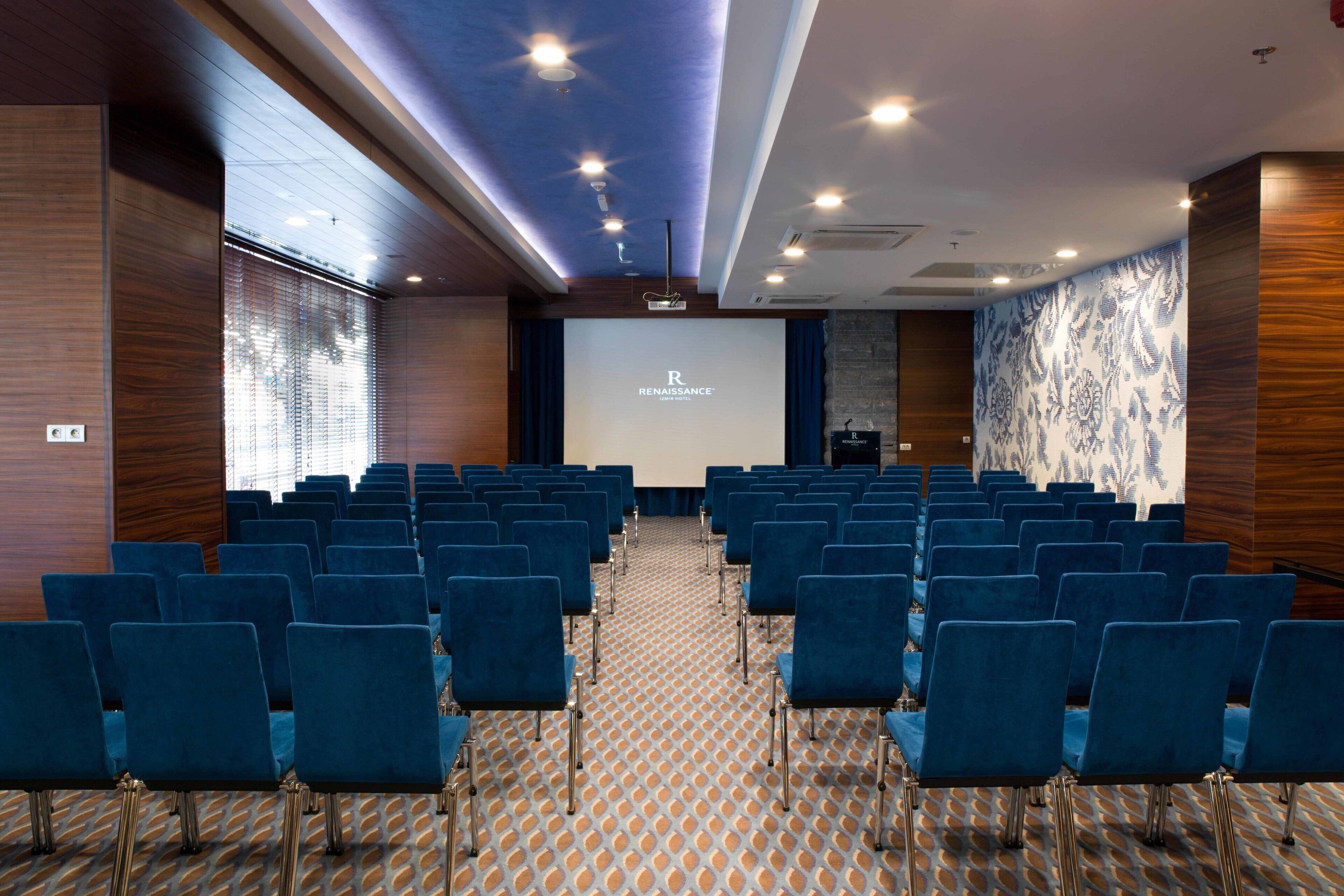 Un salón de conferencias con filas de sillas azules, una alfombra estampada, paredes de madera y una pantalla grande al frente.