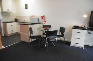 Una kitchenette compacta con microondas, un refrigerador pequeño y una cafetera. Hay una mesa de vidrio con cuatro sillas negras y un televisor pequeño sobre un mueble.