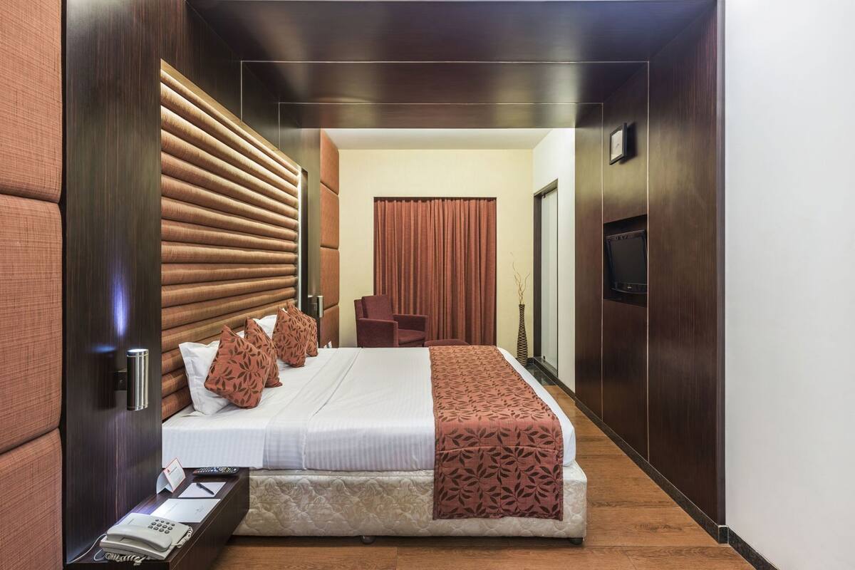 A hotel room with a bed, a desk with a phone, a chair, a TV, and a large window with curtains.