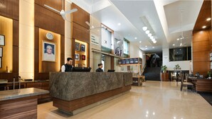 A modern hotel lobby with a reception desk, wooden paneling, a clock, framed pictures, and a staircase leading to the upper floor. - GCC Hotel and Club (Mumbai)