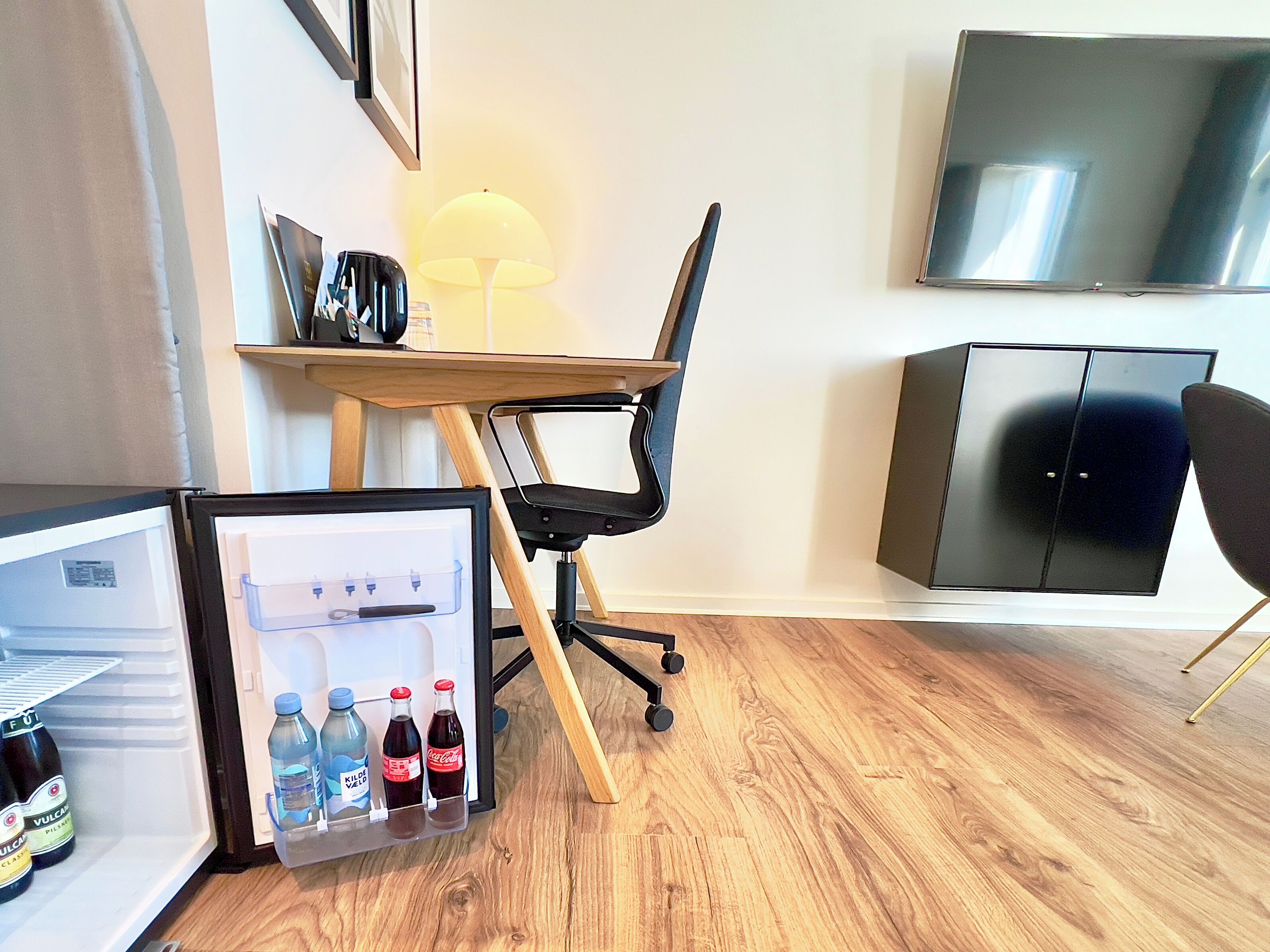 A compact room with a wooden floor, a desk with a chair, a TV mounted on the wall, a black cabinet, and a mini-fridge with beverages.