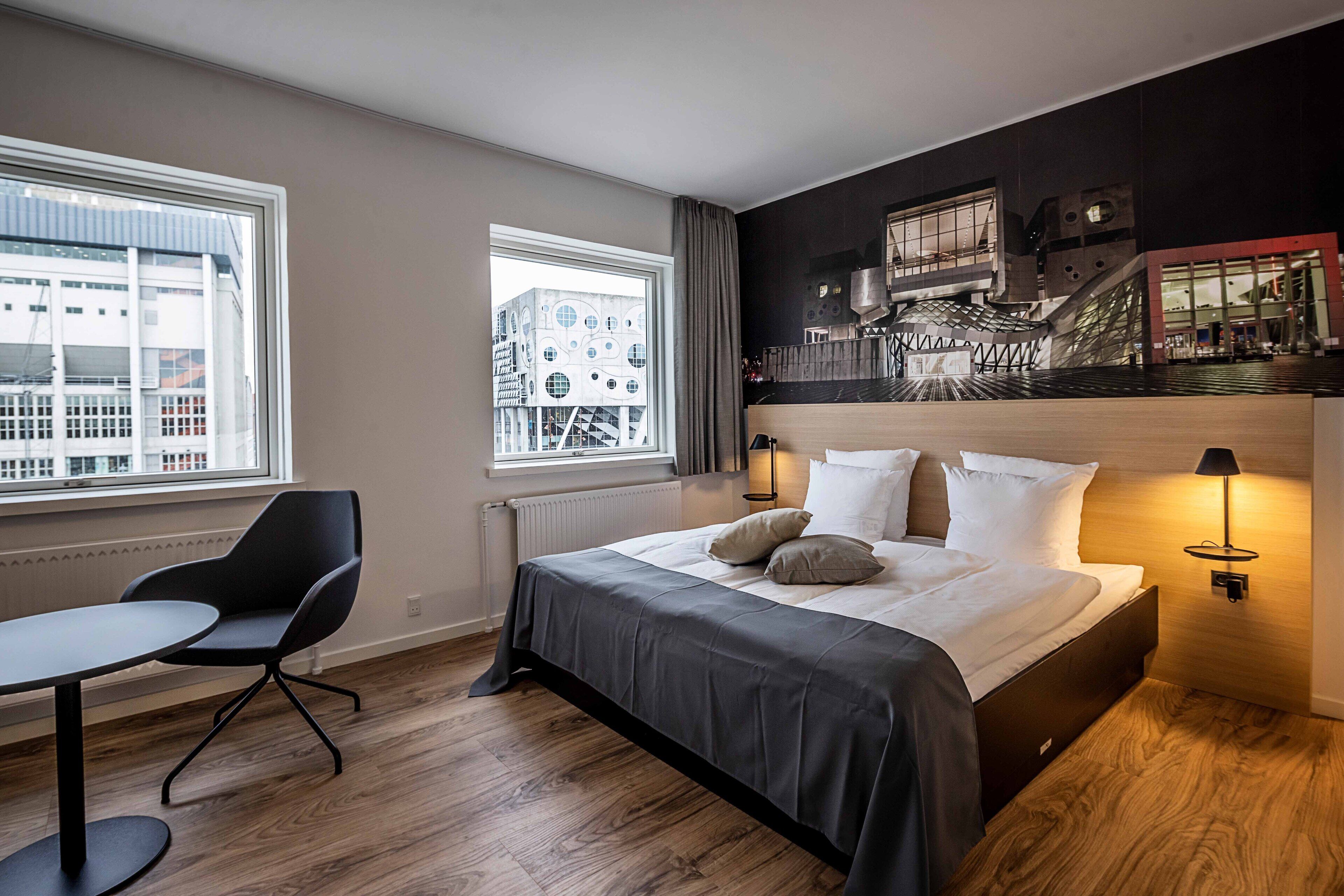 A modern hotel room with a large bed, a desk, and a chair. There are two windows, one with a view of a building and another with a view of a bridge.