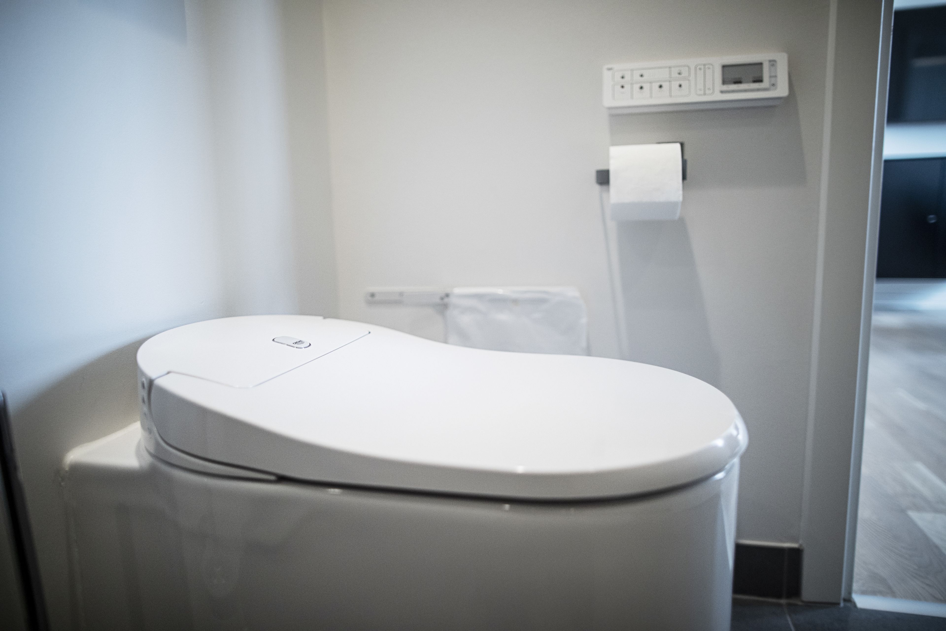 Un baño moderno con inodoro blanco, dispensador de papel higiénico en la pared y un enchufe visible.