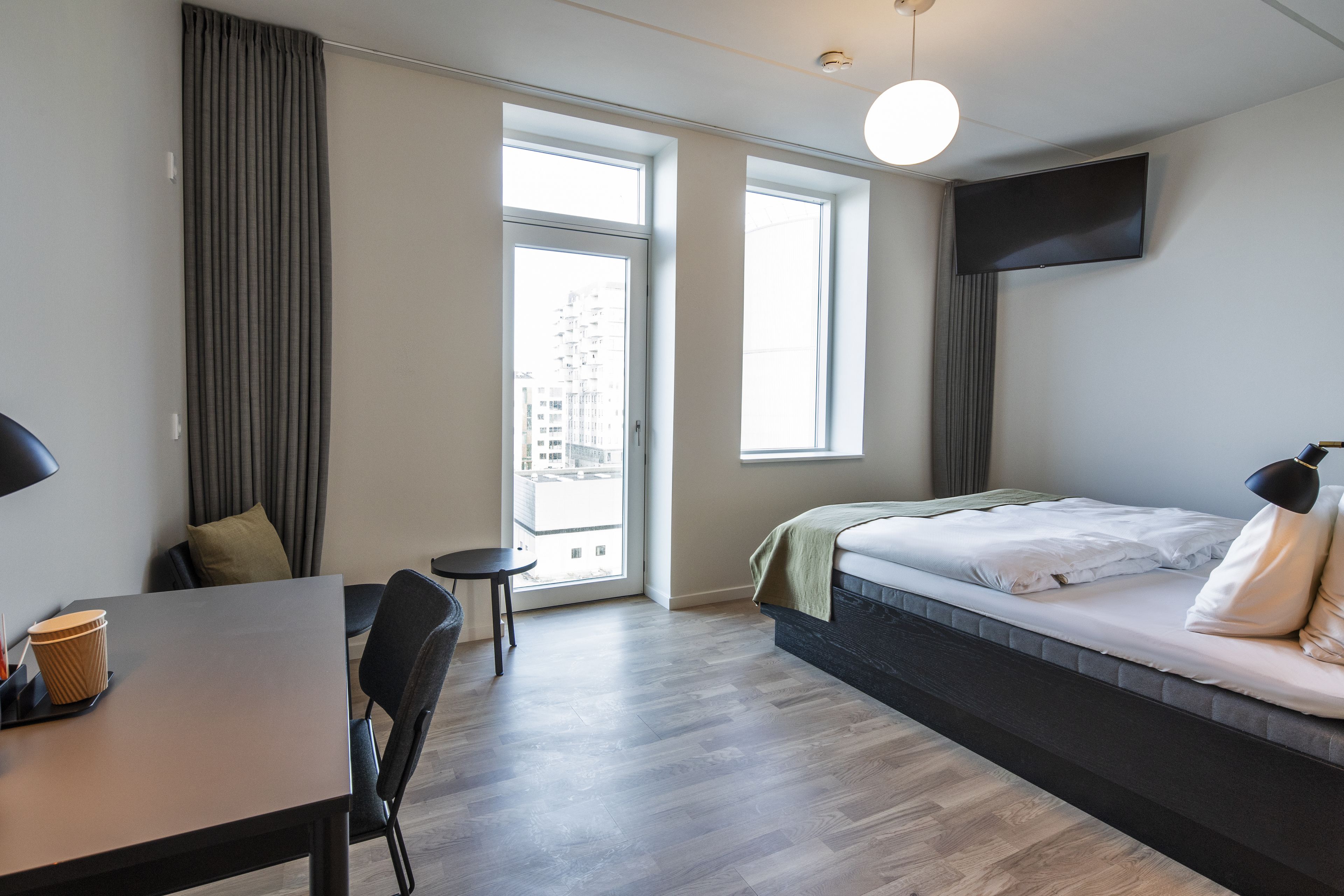 A modern hotel room with a bed, a desk, a chair, and a large window offering a city view.