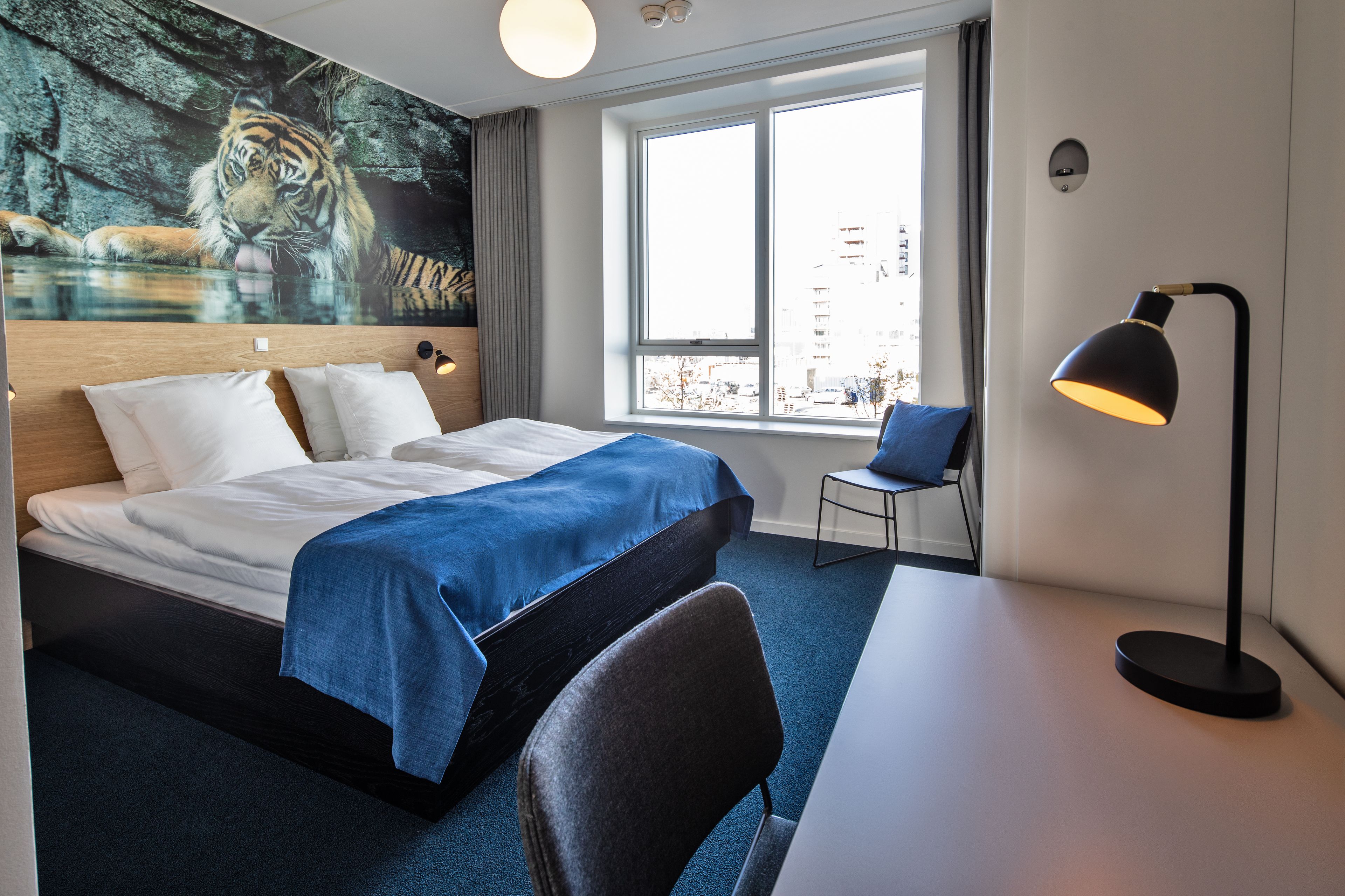 Et hotelværelse med en seng, et skrivebord, en stol, et vindue med udsigt til bygninger og et tiger-maleri på væggen.