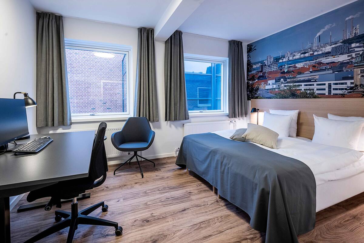 A hotel room with a bed, a desk with a computer, a chair, and a large window with curtains.