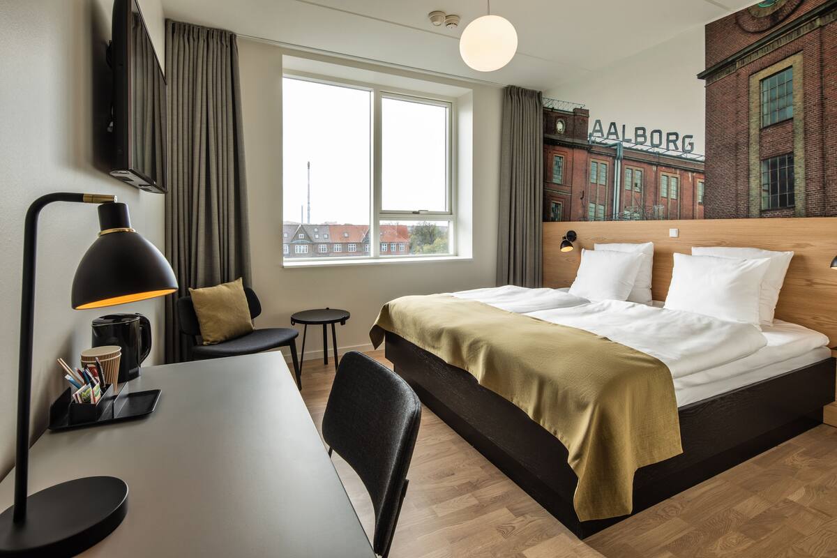 A modern hotel room with a large bed, a desk, a chair, a lamp, and a view of a building outside.