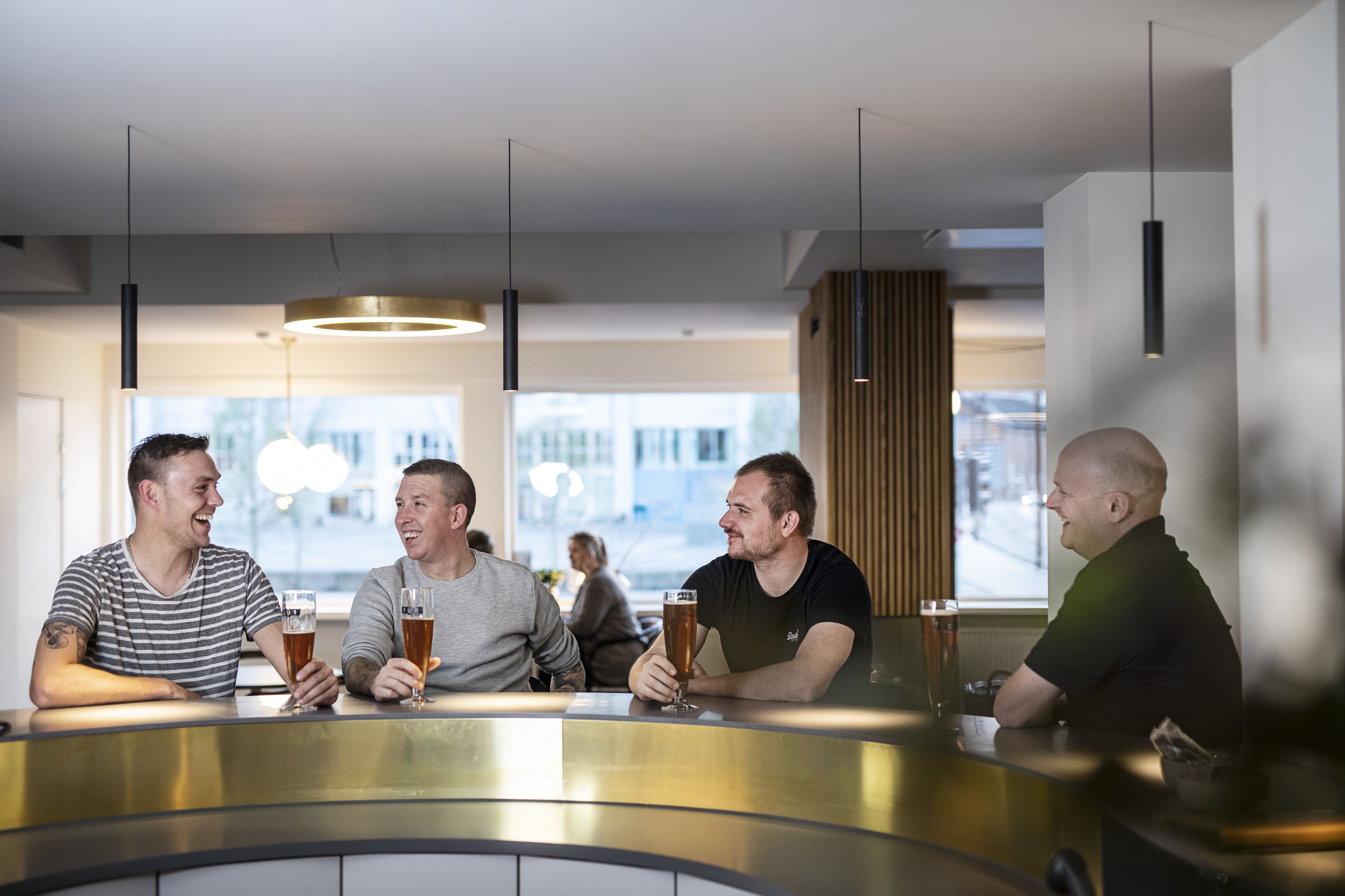 Un grupo de personas sentadas en un bar con cervezas, en un interior moderno con iluminación colgante y ventanas amplias.