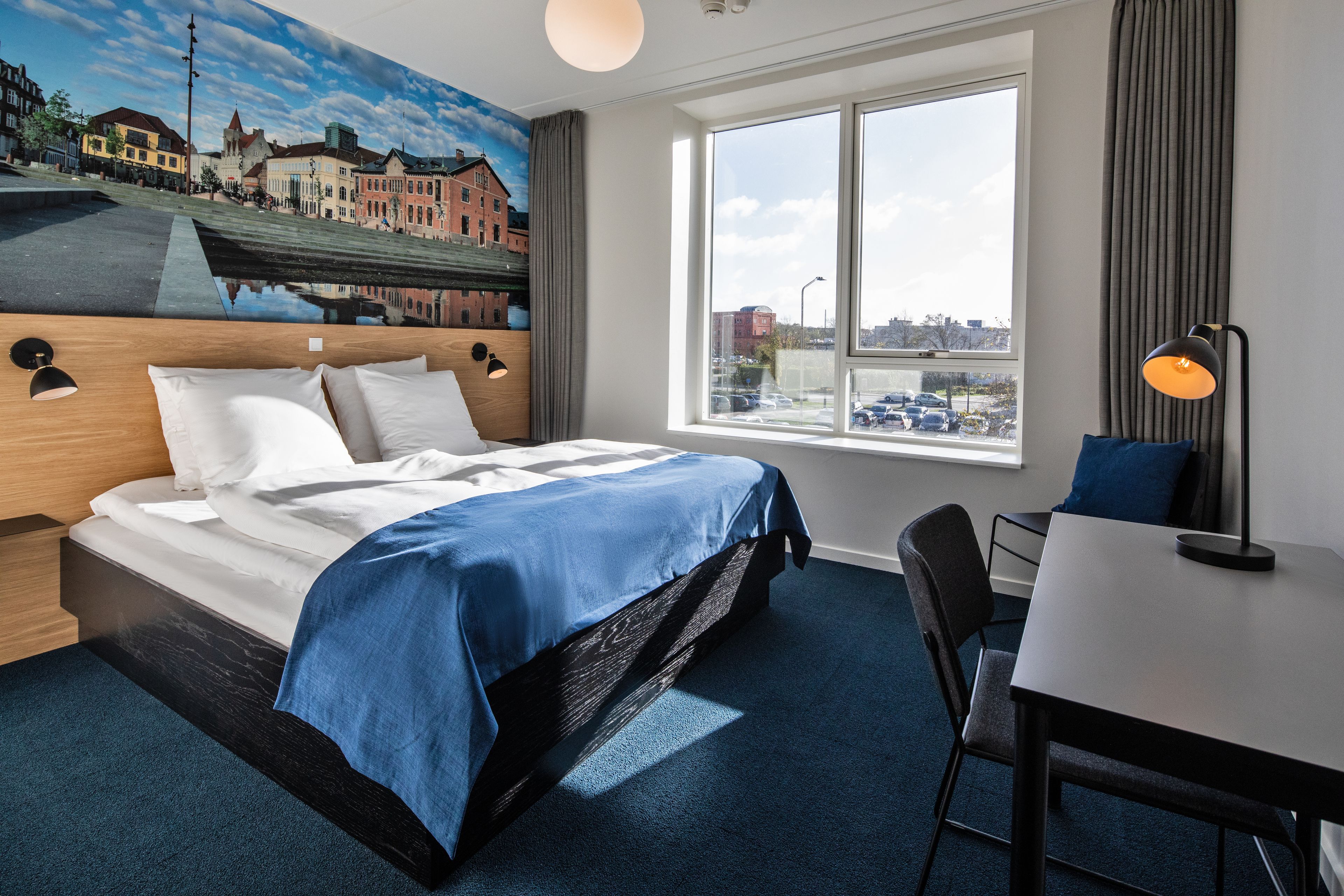 A hotel room with a bed, a desk, a chair, a large window with a view of buildings and a canal, and a mural on the wall.