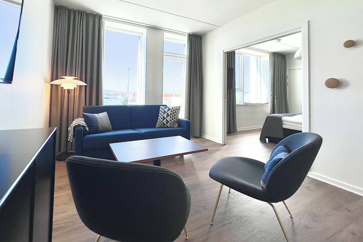 A modern hotel room with a blue sofa, a small table, two chairs, and large windows offering a view of the city.