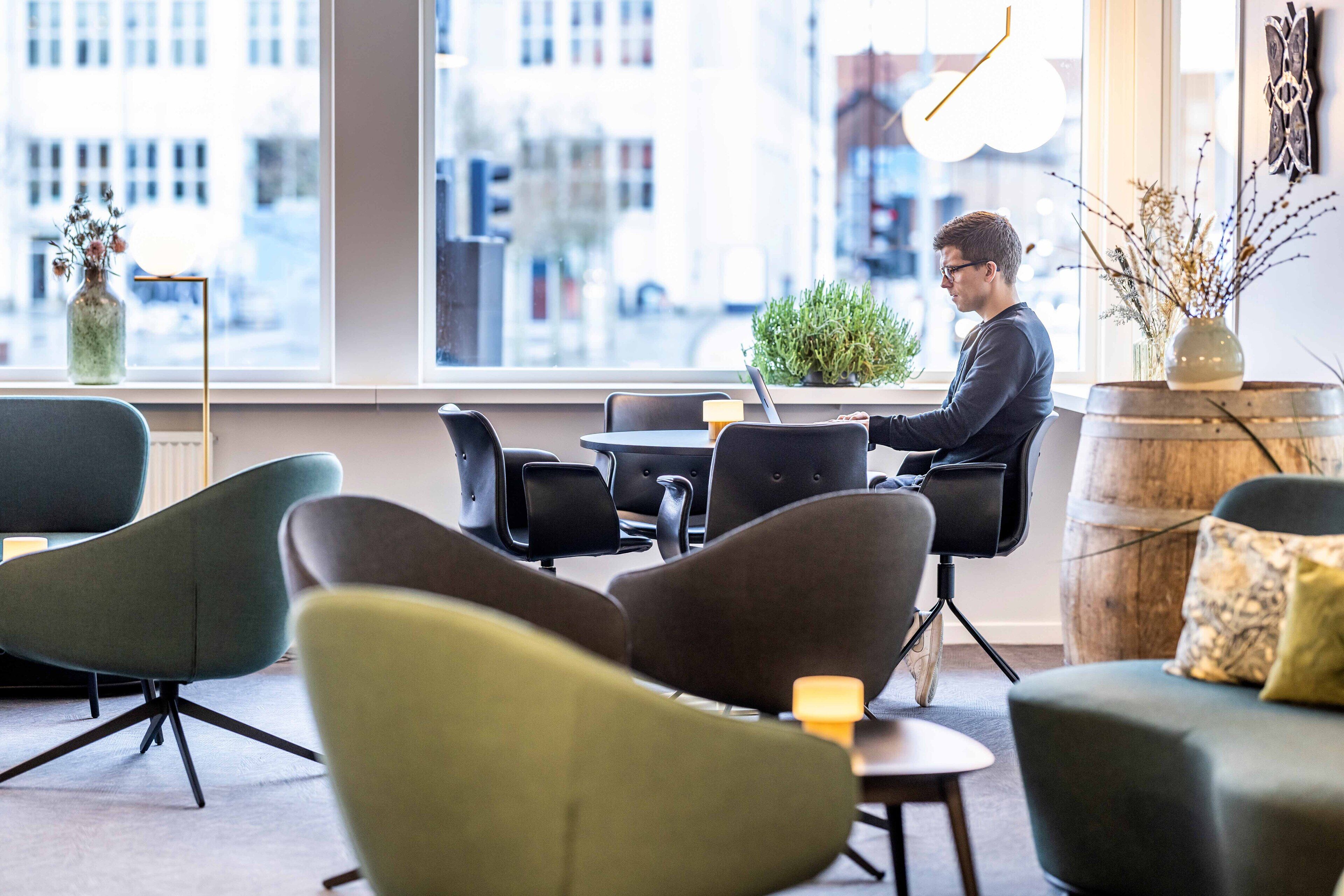A modern interior with a man working at a laptop, surrounded by comfortable seating and a large window offering a city view.