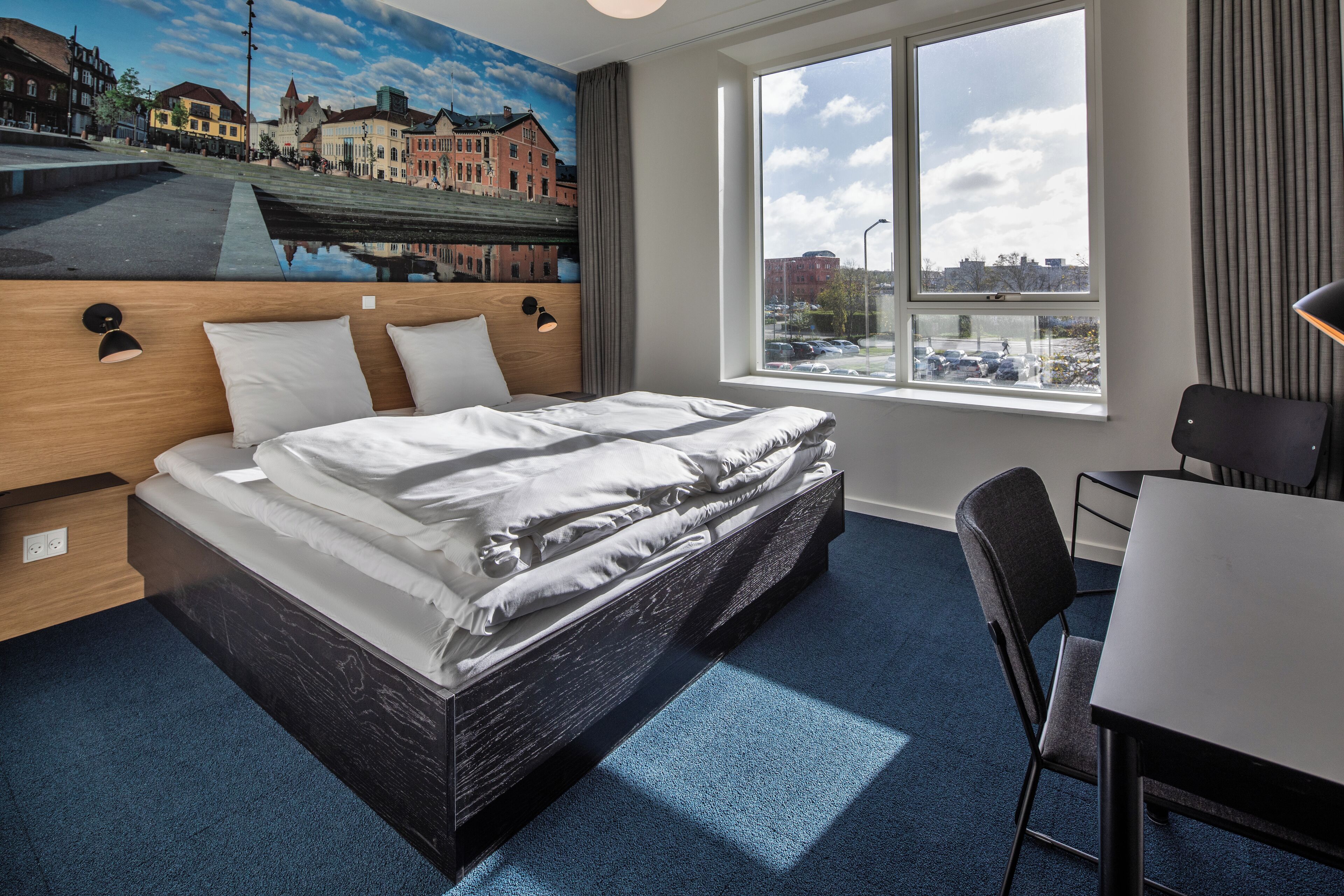 A hotel room with a bed, a desk, a chair, a large window with a city view, and a mural on the wall.