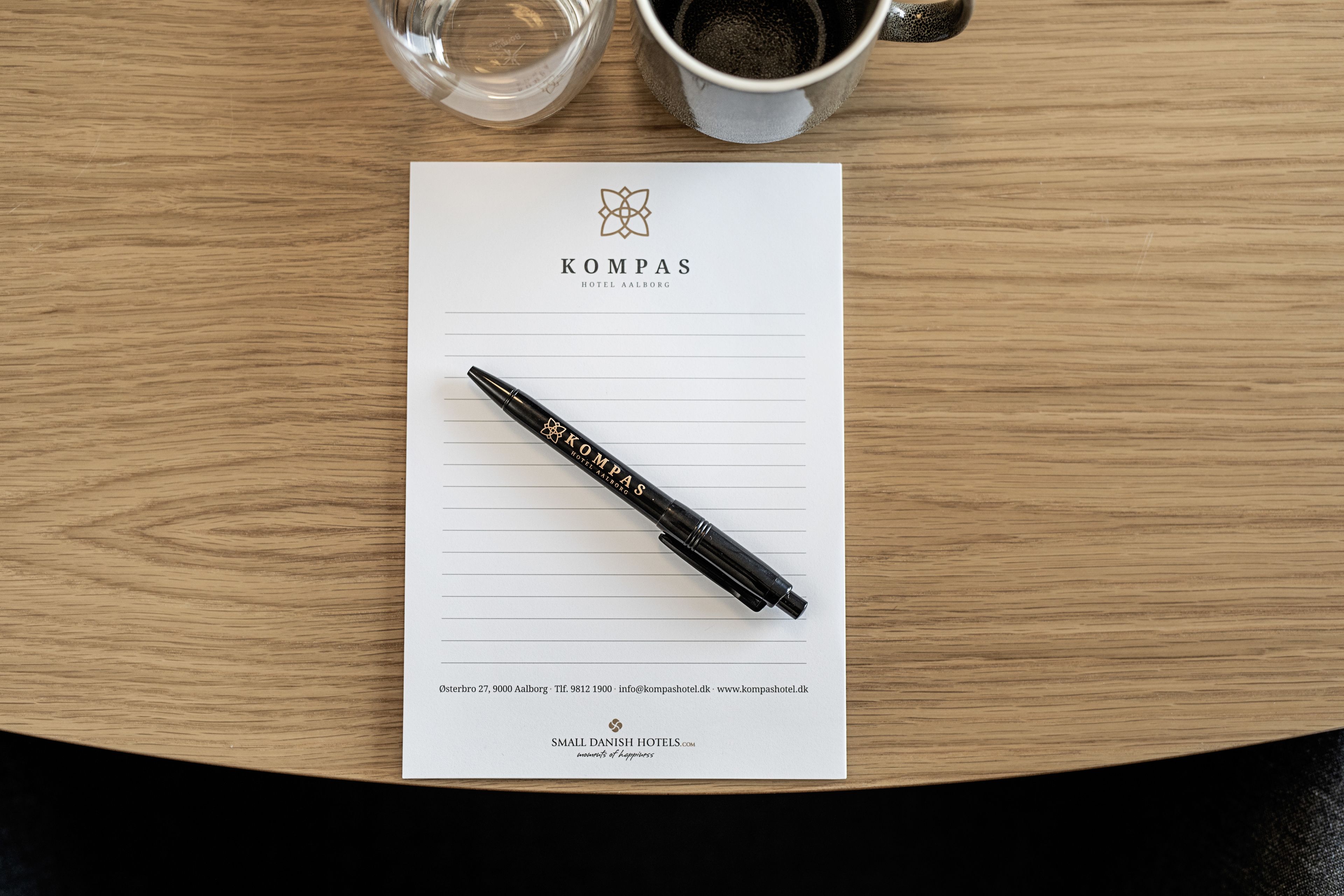 Un folleto de hotel con el logotipo de KOMPAS, un bolígrafo y una taza de café sobre una mesa de madera.