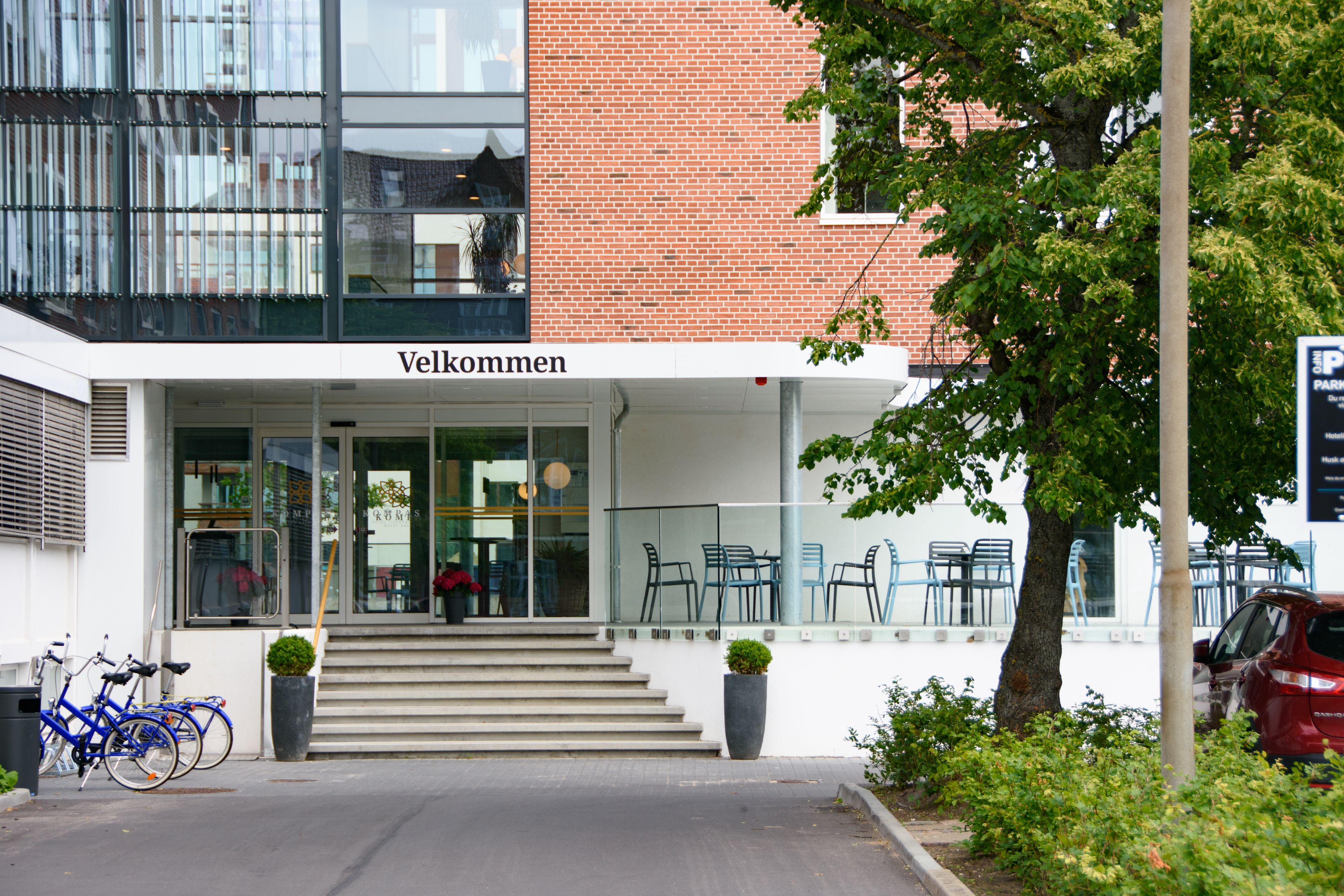 A building entrance with a sign reading "Vellkommen" and a staircase leading to the entrance. There are blue chairs outside and a red car parked nearby.