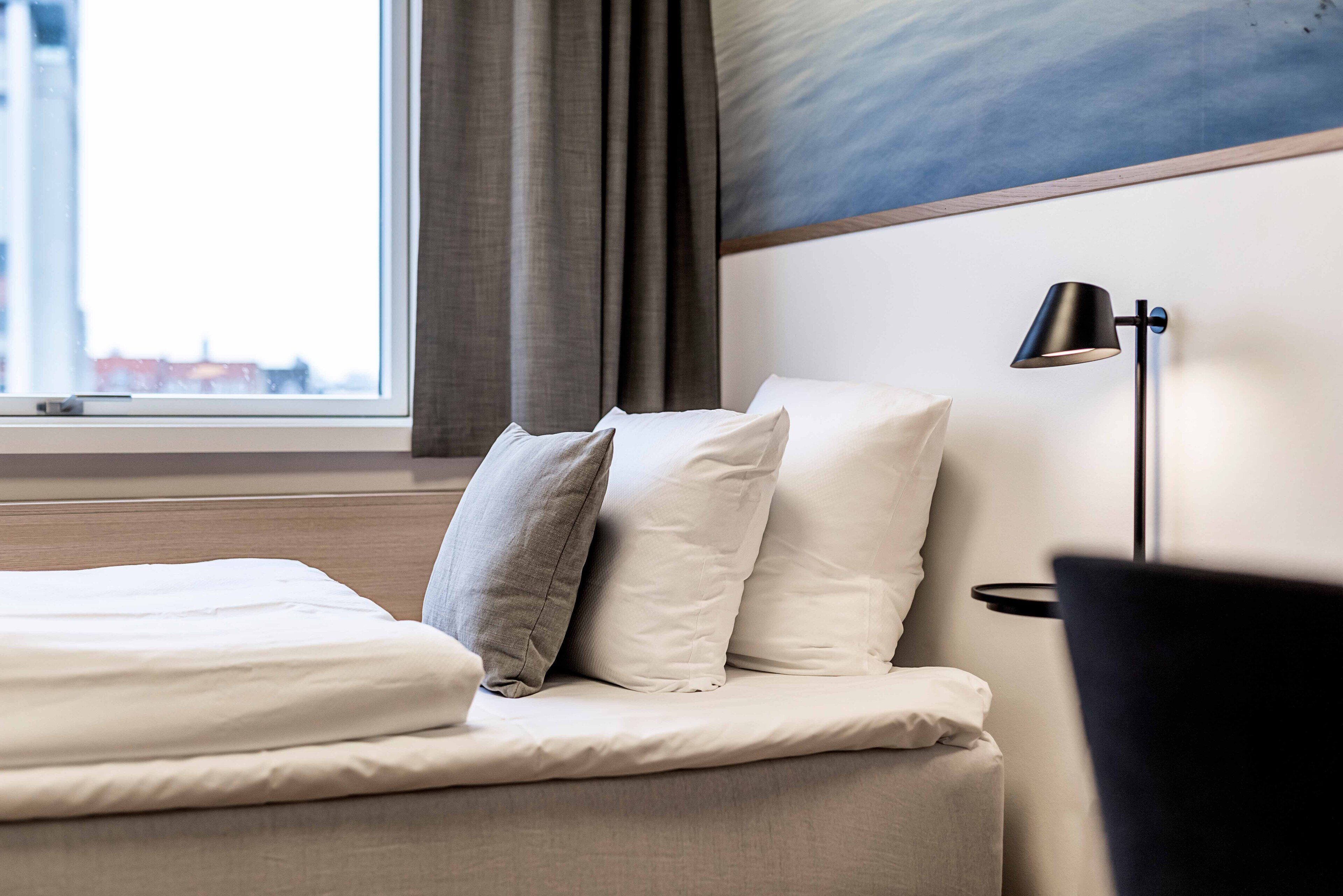 Una habitación de hotel con cama, almohadas, lámpara y ventana con cortinas.