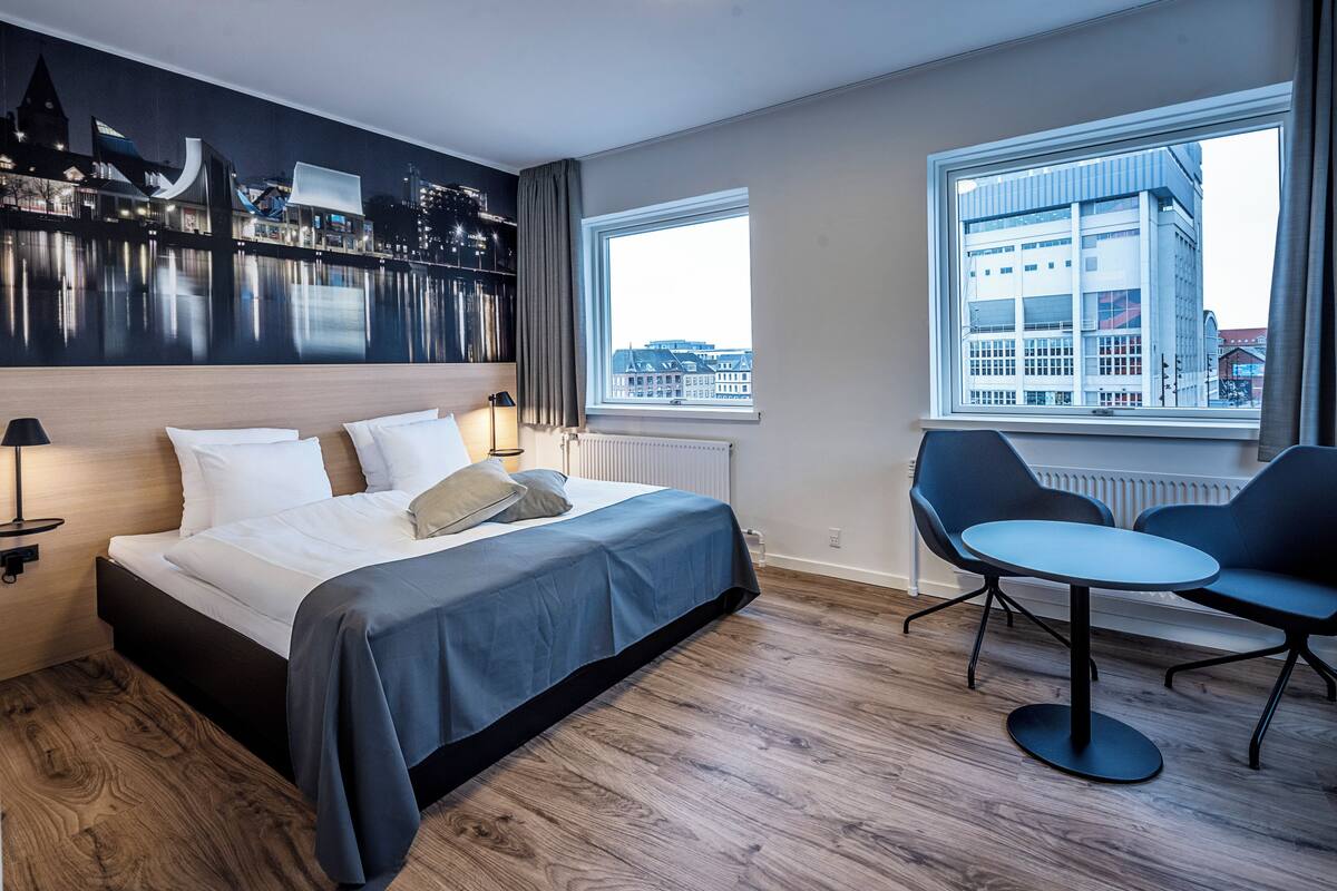 A modern hotel room with a bed, a desk, two chairs, and a view of a cityscape through large windows.