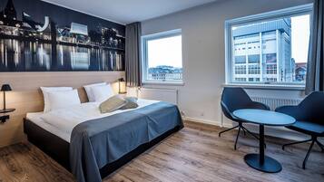 A modern hotel room with a bed, a desk, two chairs, and a view of a cityscape through large windows.