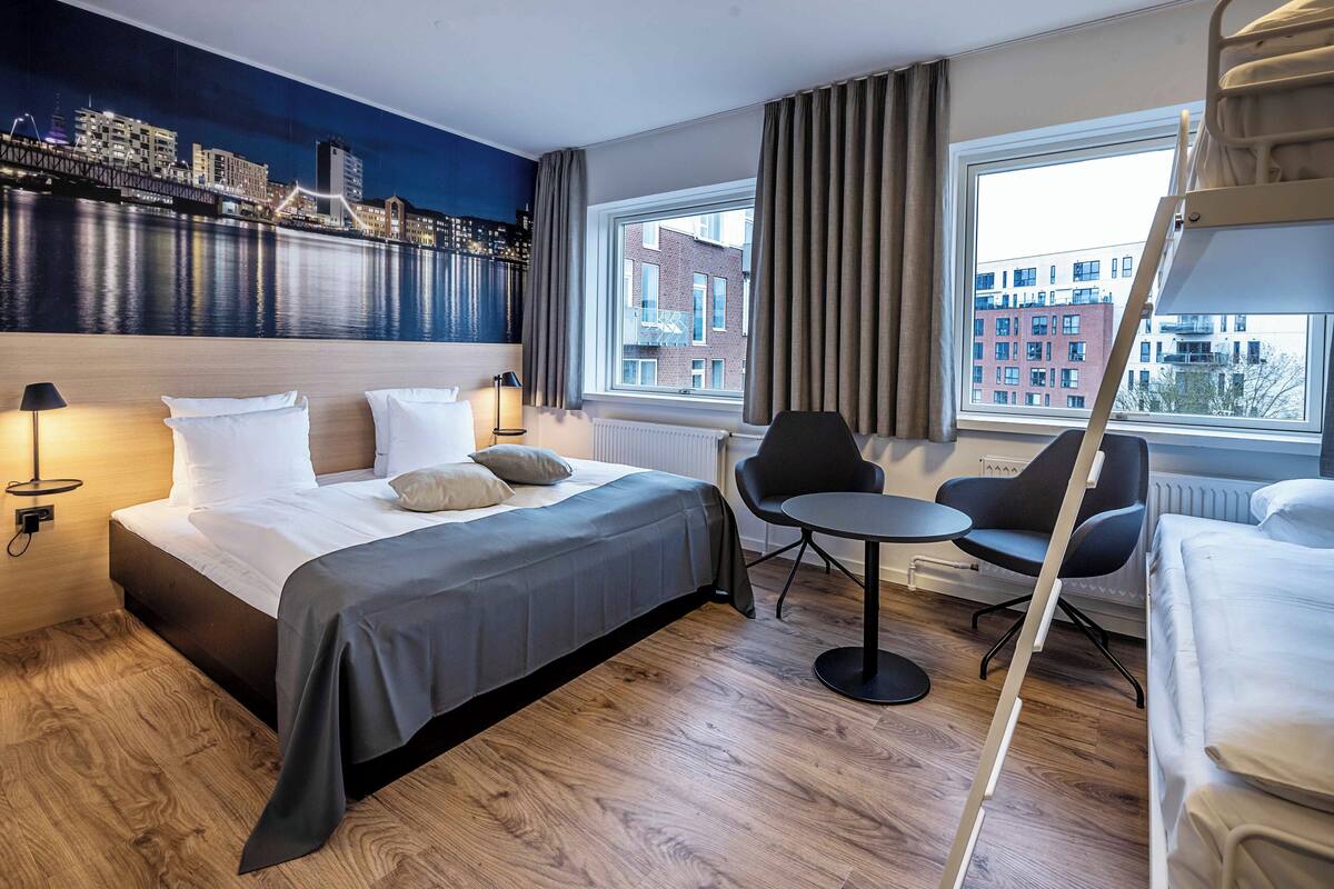 A modern hotel room with a large bed, a small round table, two chairs, a view of the city through a window, and a cityscape mural on the wall.