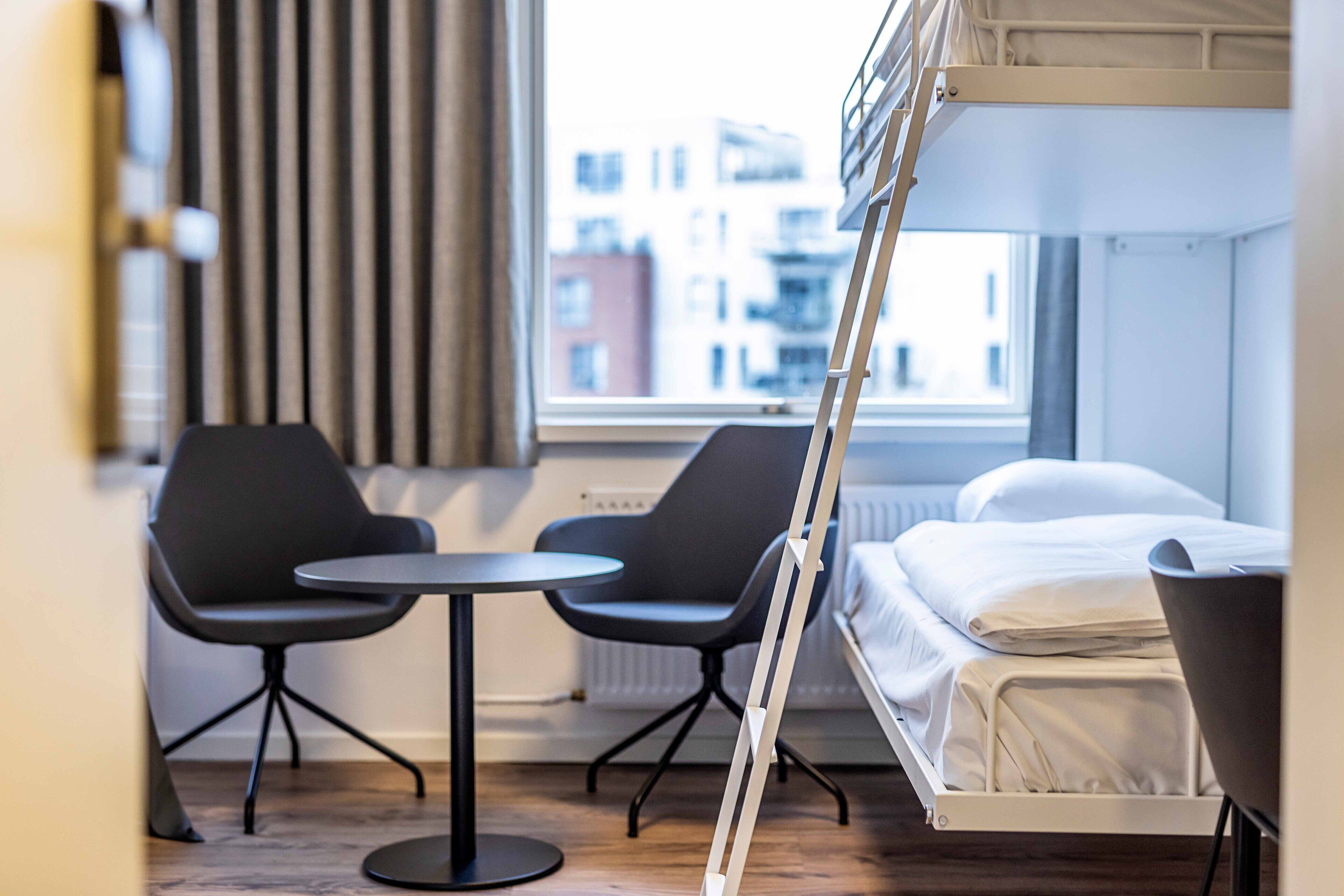 Ein kleines Hotelzimmer mit Etagenbett, zwei schwarzen Stühlen, einem runden Tisch und einem Fenster mit Blick auf die Stadt.