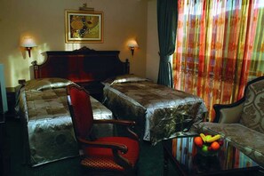 A hotel room with two beds, a red chair, a table with fruit, and a painting on the wall. - Millenium Palace Bitola (Bitola)