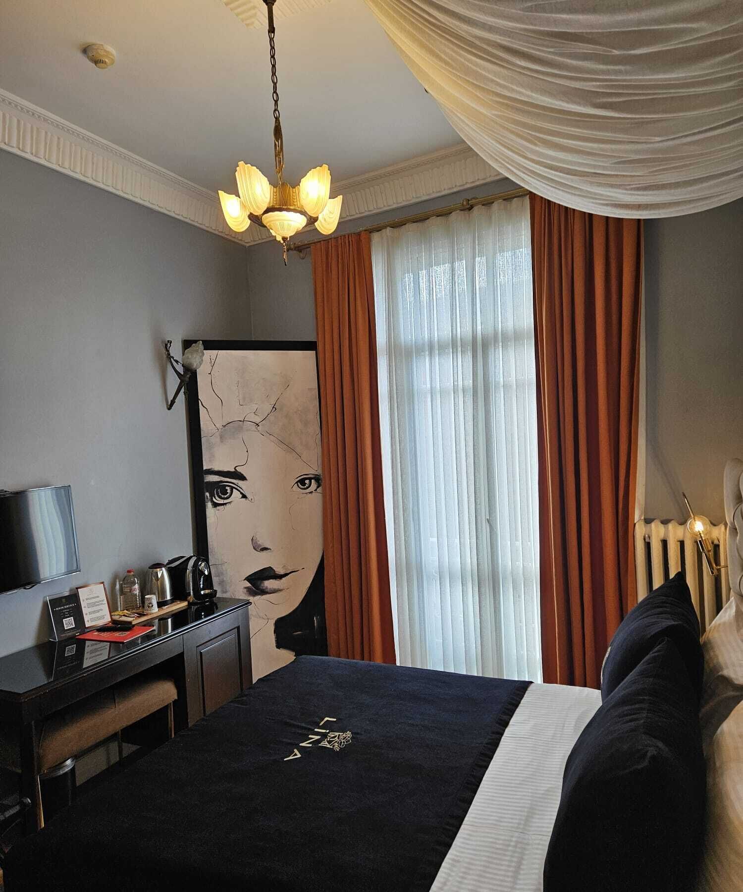 A hotel room with a bed, a desk with a computer, a chair, a lamp, and a large window with curtains.