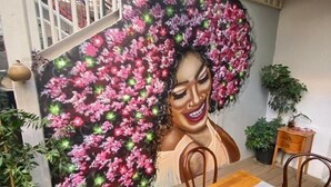 Un comedor con un mural de una mujer rodeada de flores, sillas de madera, una mesa con mantel blanco y plantas en macetas.
