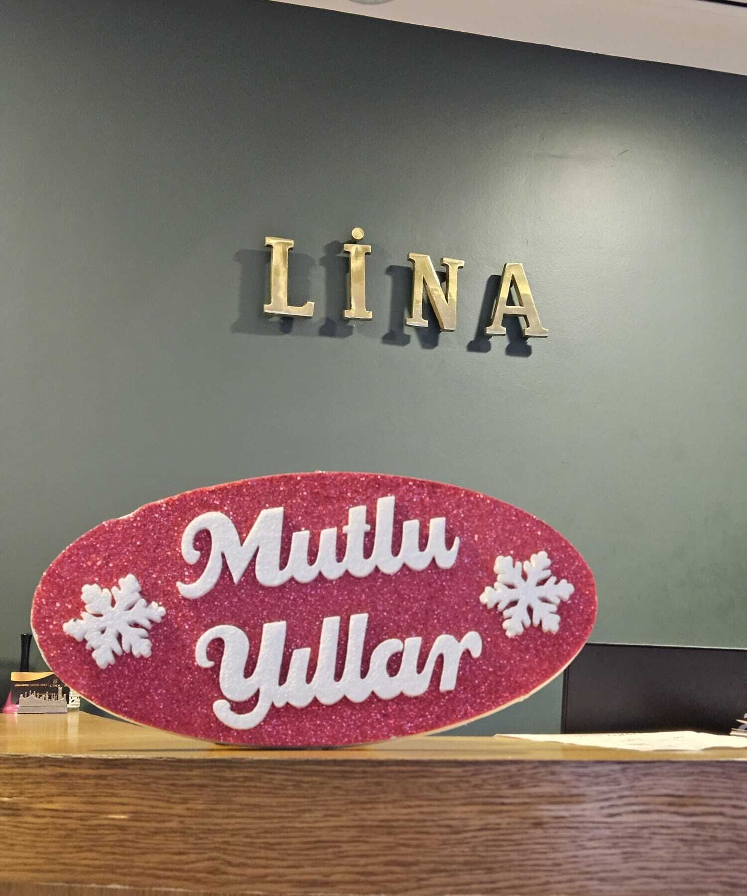 Een receptiebalie met een bord waarop 'LİNA' staat en een decoratief bord eronder met de tekst 'Mutlu Yillar'.