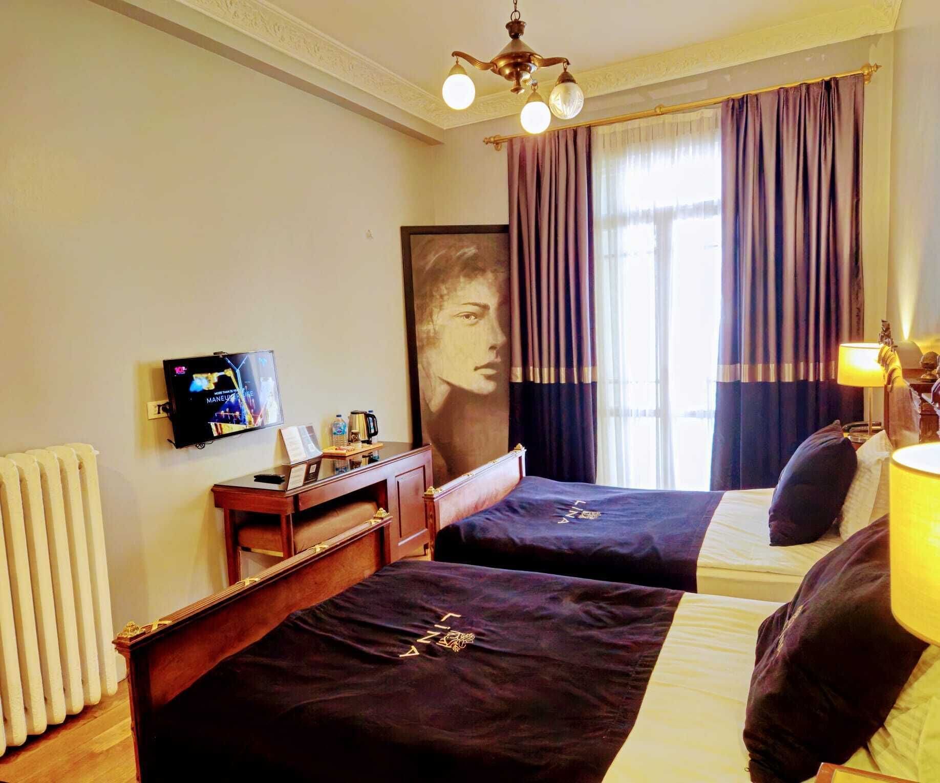 Habitación de hotel con dos camas, televisión, radiador y un amplio ventanal con cortinas.
