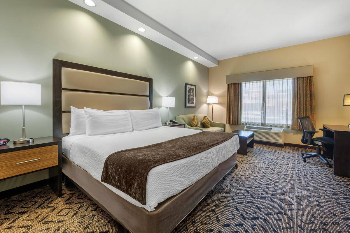 Een hotelkamer met een groot bed, nachtkastjes, een bureau, een stoel, een lamp, een schilderij aan de muur en een raam met gordijnen.
