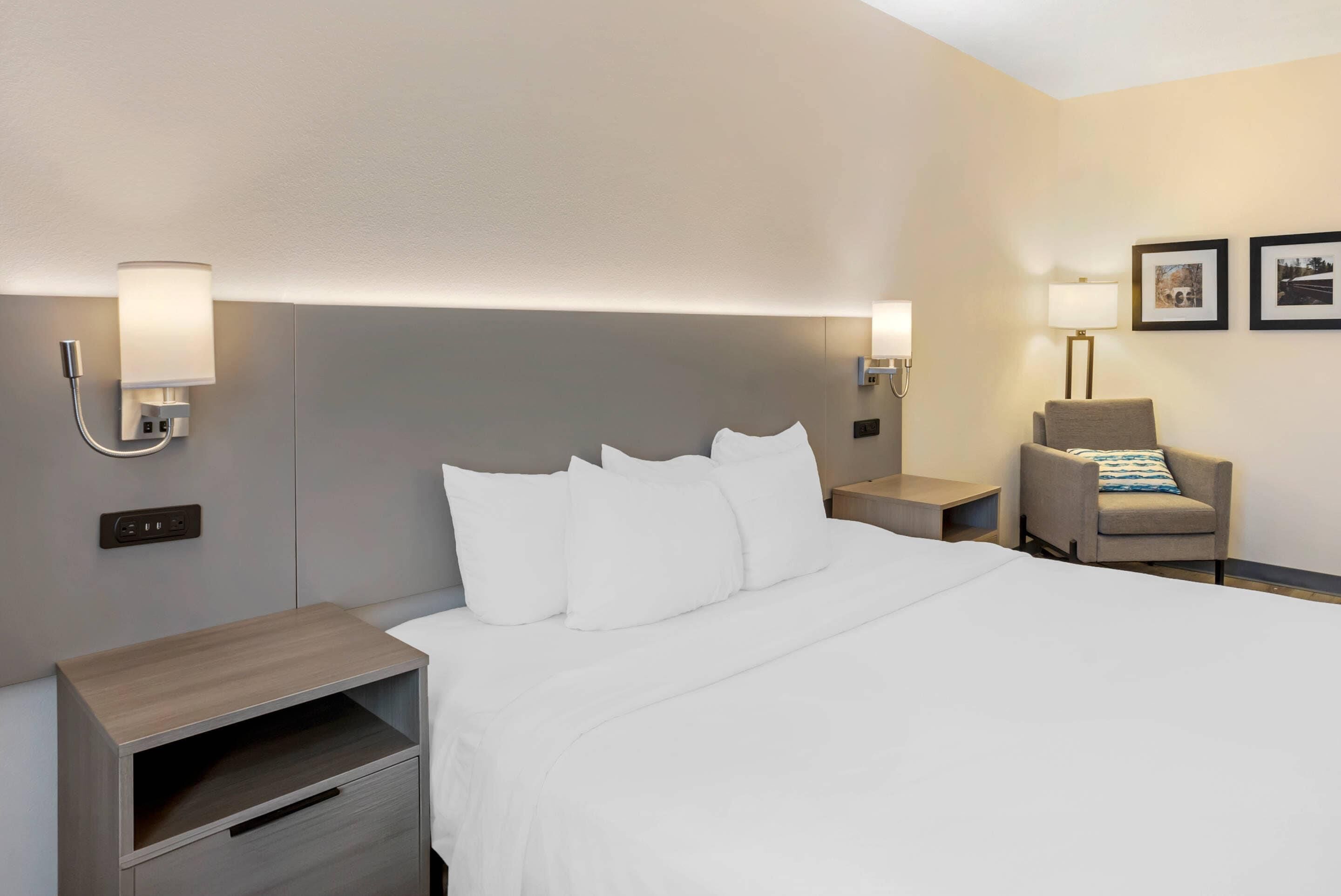 Una habitación de hotel con una cama grande, mesitas de noche, una silla y lámparas de pared.