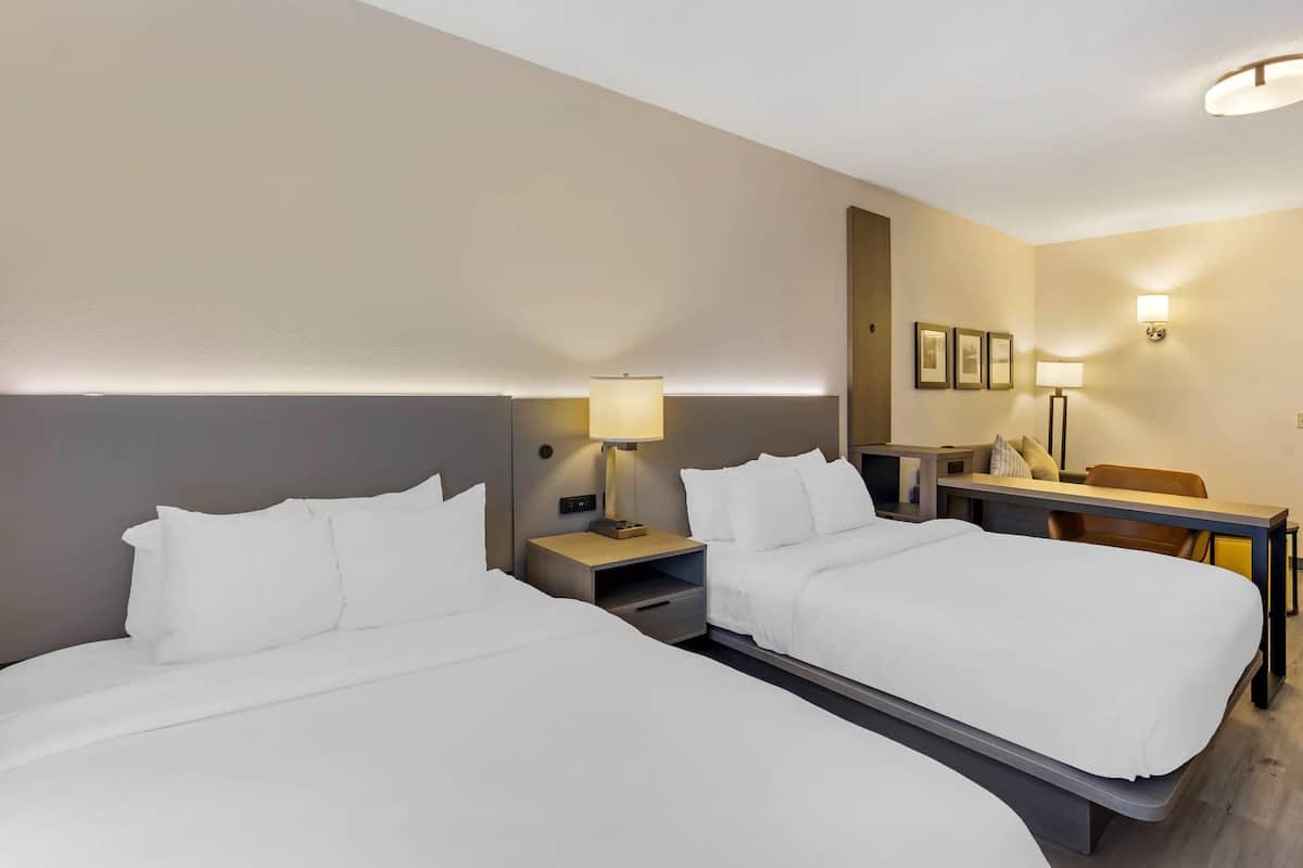 A hotel room with two beds, a desk, a chair, and a lamp.