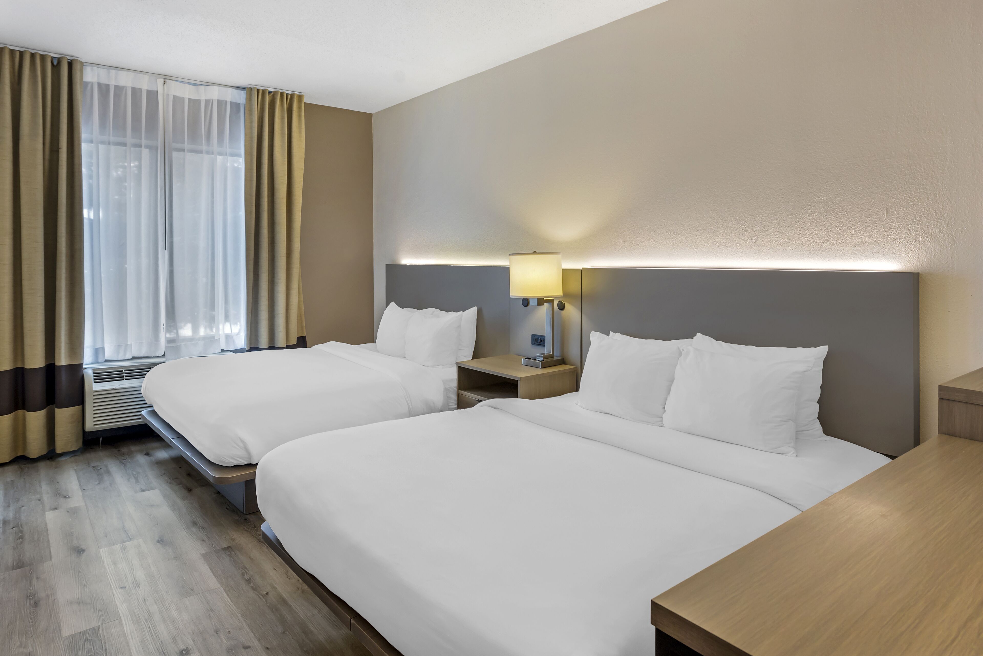 A hotel room with two beds, a bedside table with a lamp, a window with curtains, and a wooden floor.