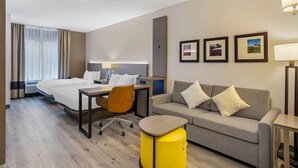 A hotel room with a bed, a desk, a chair, a sofa, and a small yellow ottoman.