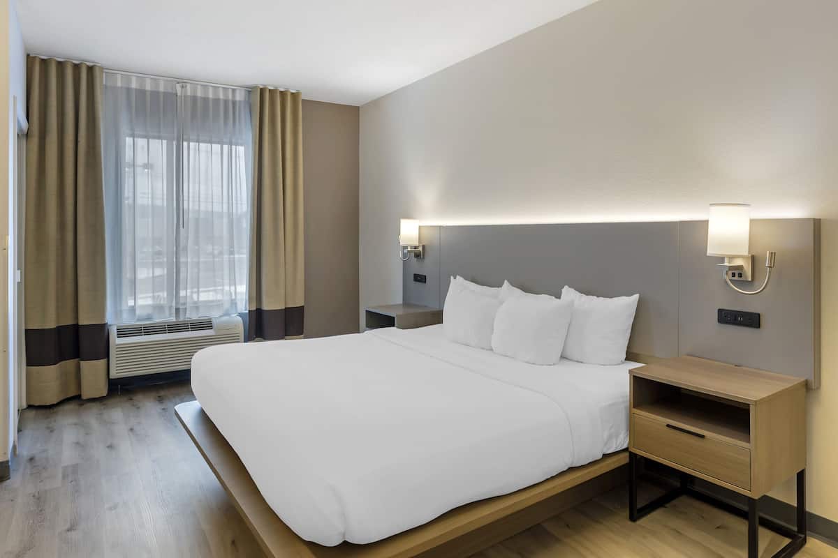 A hotel room with a large bed, bedside tables, a nightstand, a wall-mounted lamp, and a window with curtains.