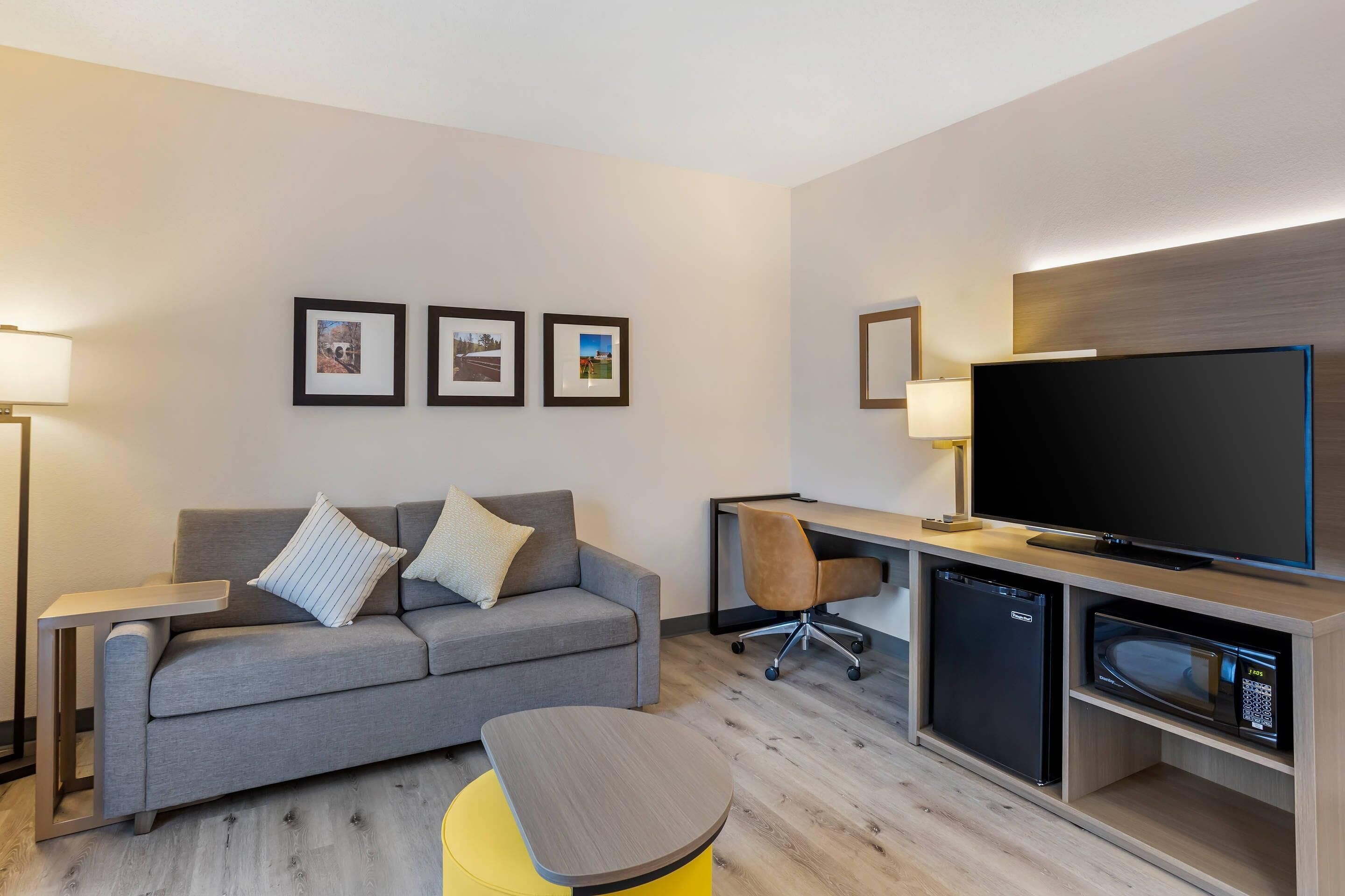 Una moderna camera d'albergo con un divano grigio, un poggiapiedi giallo, una TV a schermo piatto, un mini-frigorifero e una scrivania con una sedia.