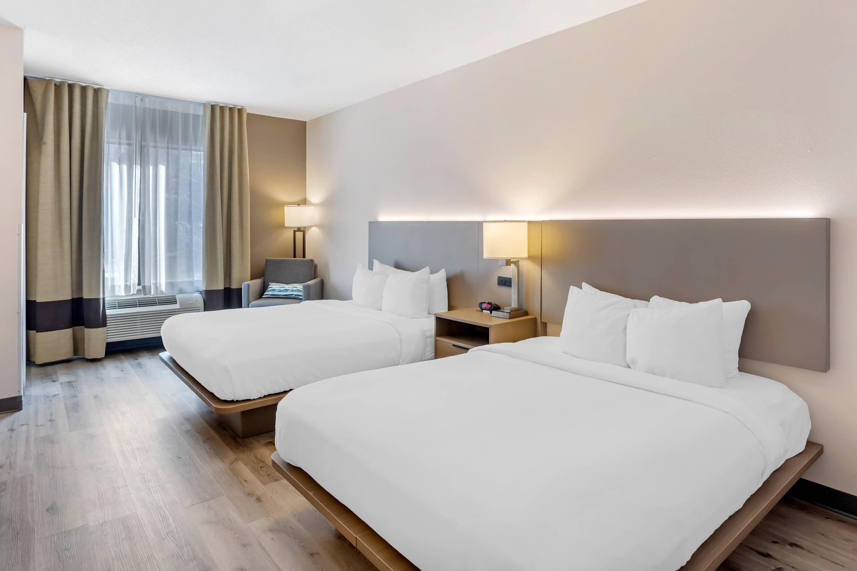 Habitación de hotel con dos camas, un escritorio, una silla y un ventanal con cortinas.